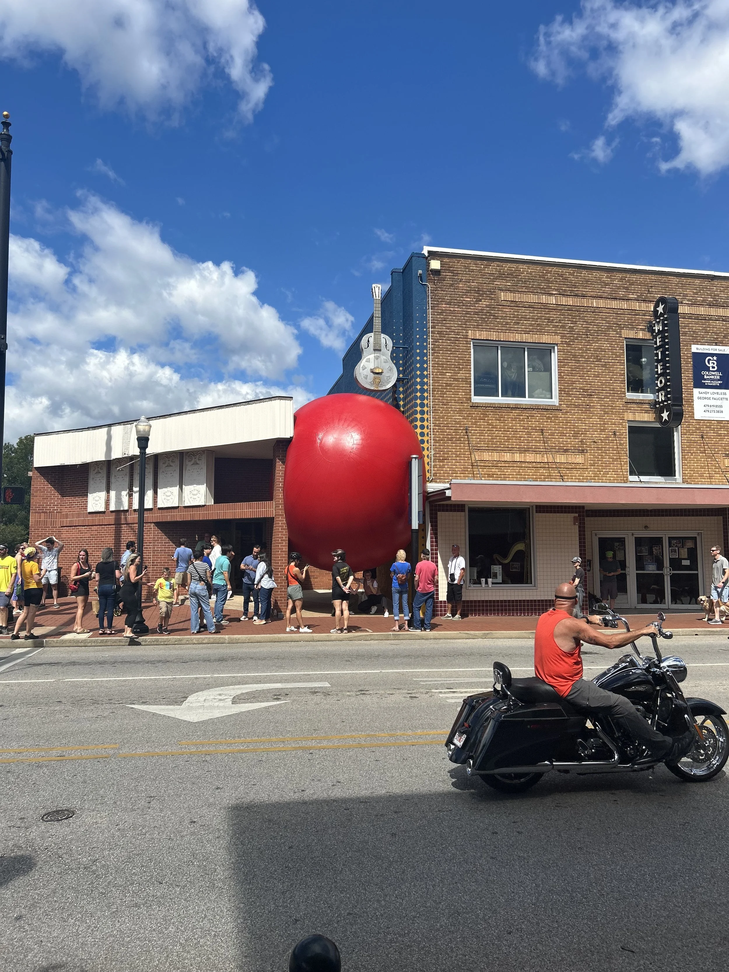 People standing in line outside a building with a large red sphere, a white guitar sign, and a small fin on top of the building. A man on a motorcycle is passing by on the street.
