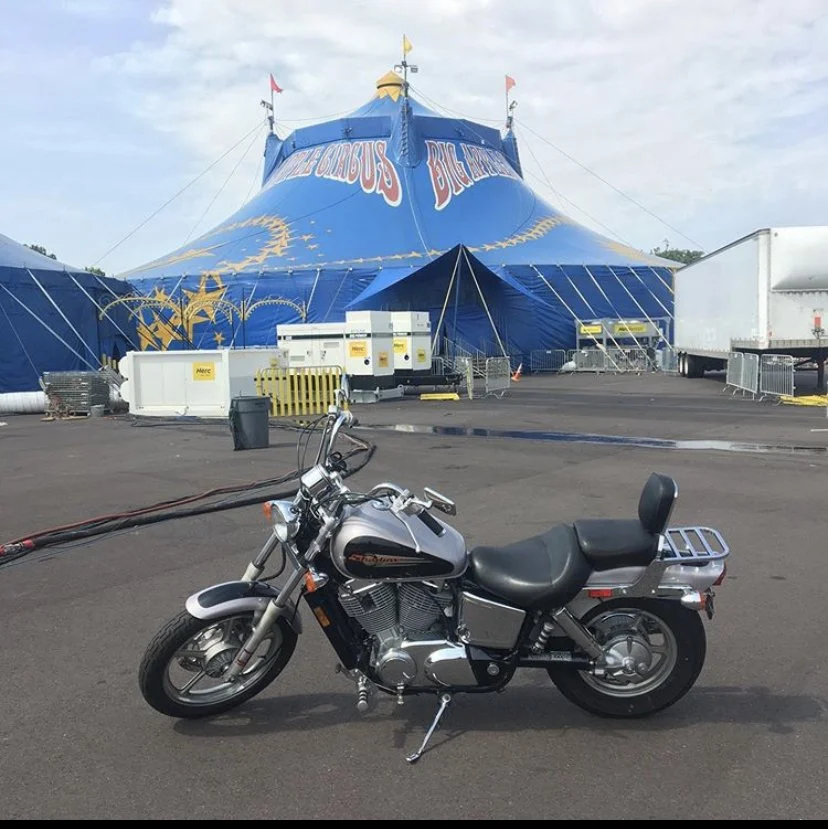 Motorcycle parked outside a big blue circus tent with red and white lettering.