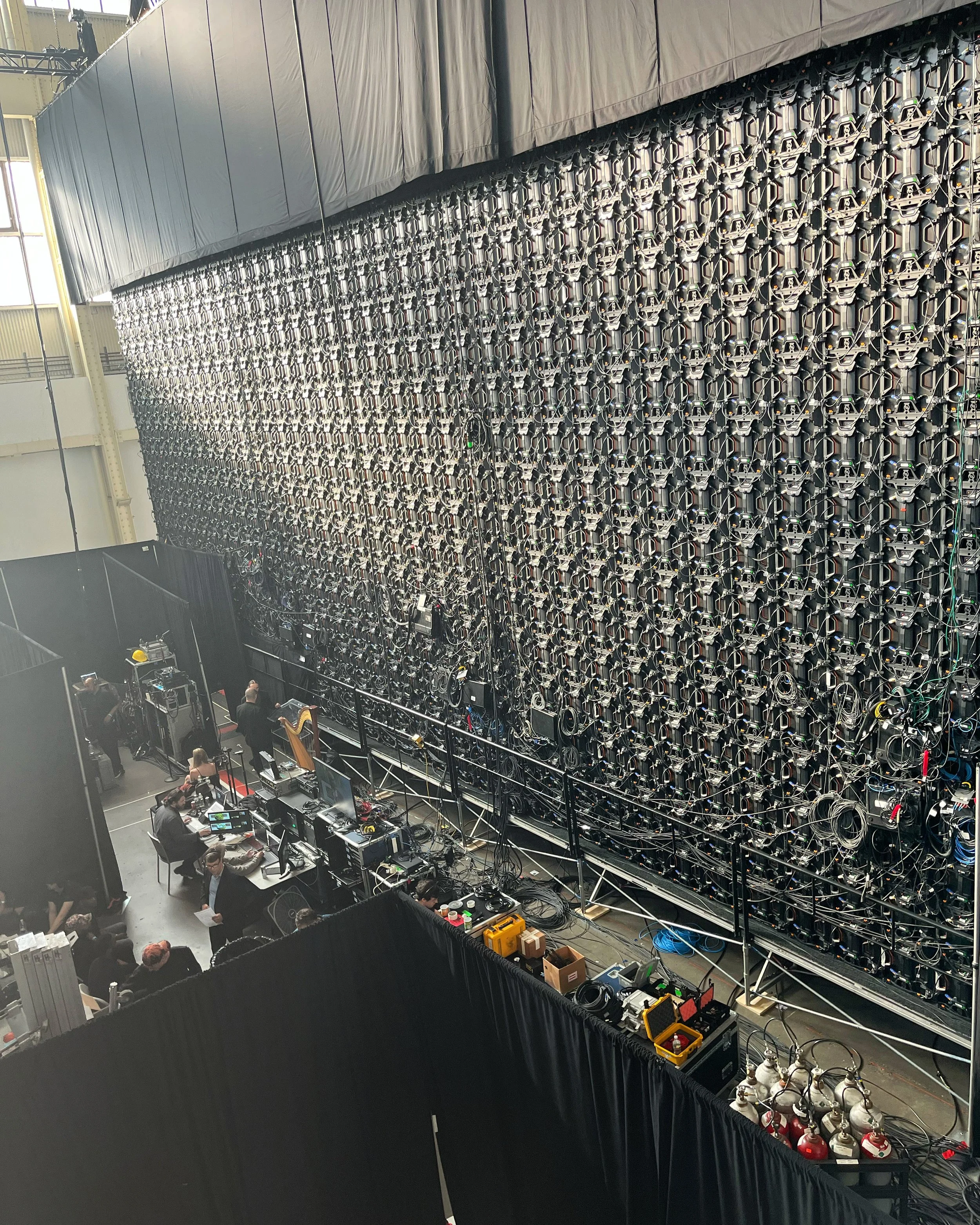 Inside a large facility with a wall of numerous synchronized lighting and electronic equipment, and technicians working at desks and equipment stations.