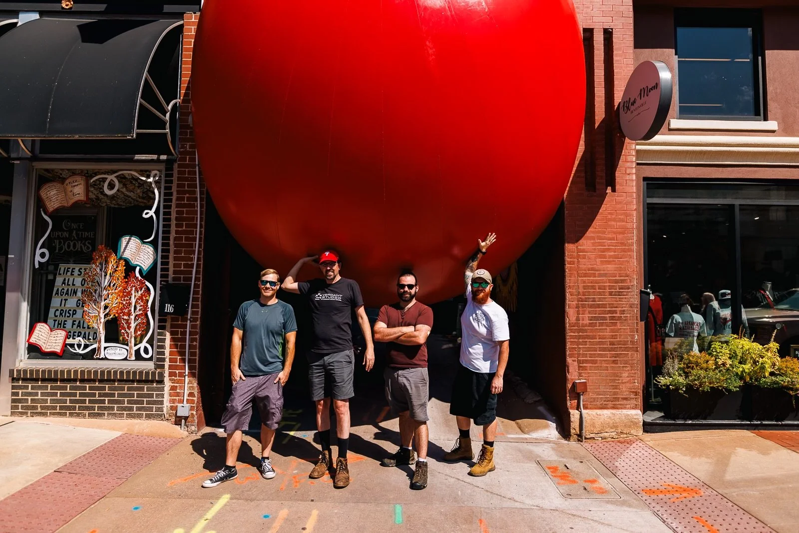 Four men standing in front of a large red apple sculpture outside a bookstore on a city sidewalk.