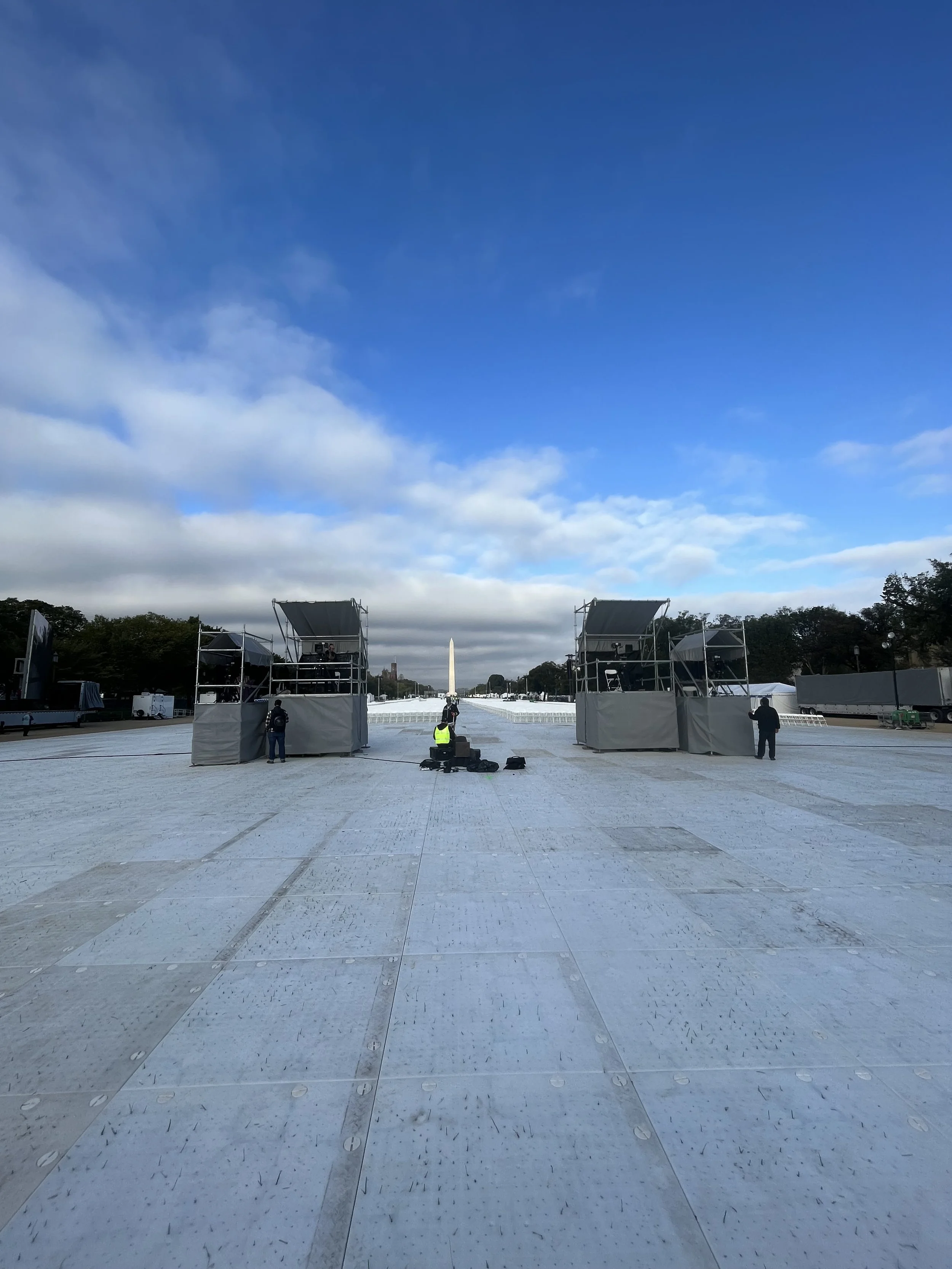 Construction workers set up stages on the National Mall with the Washington Monument in the background under a partly cloudy sky.