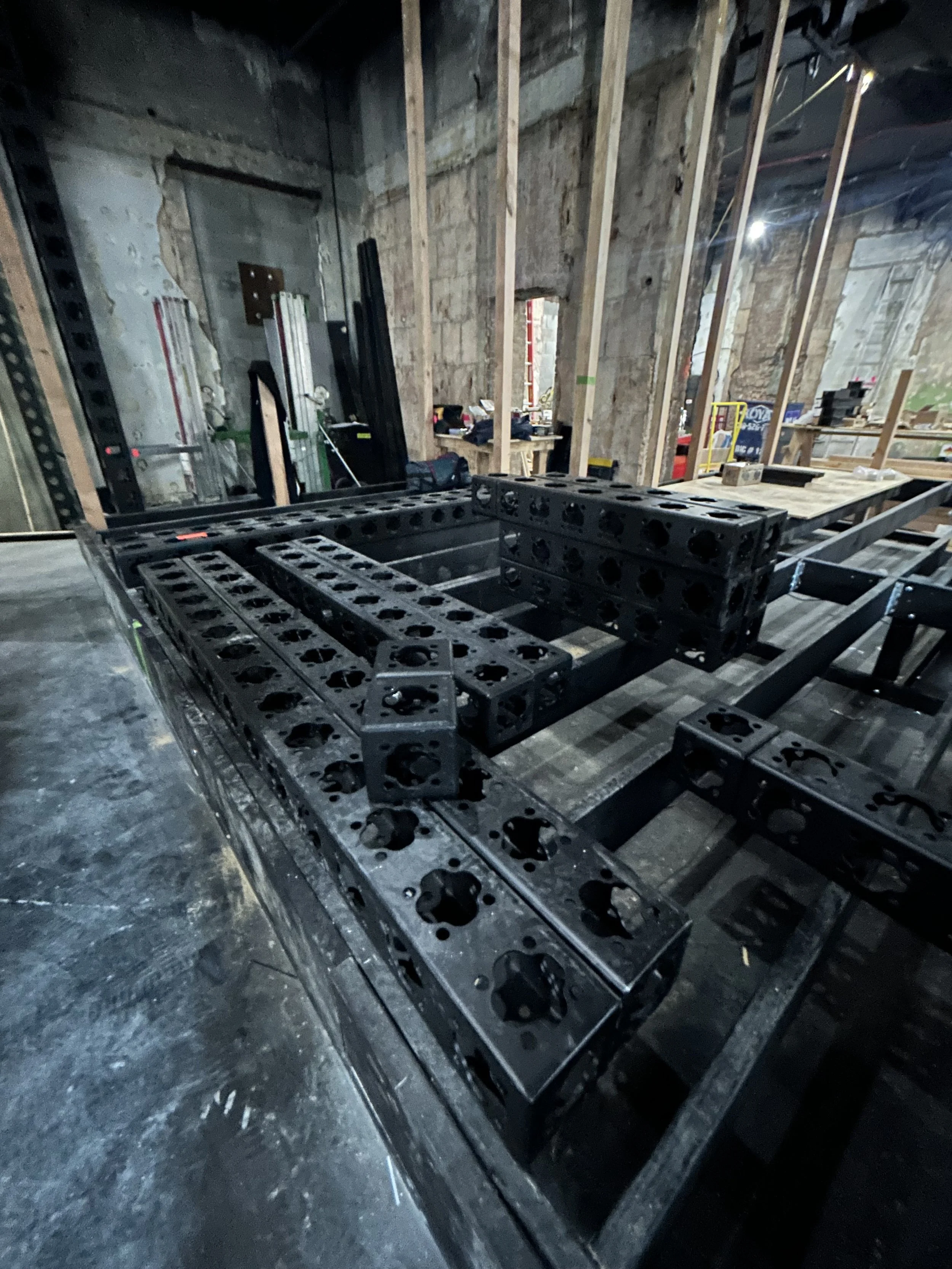 Construction site with black metal parts assembled on a wooden table, surrounded by unfinished walls and construction tools.