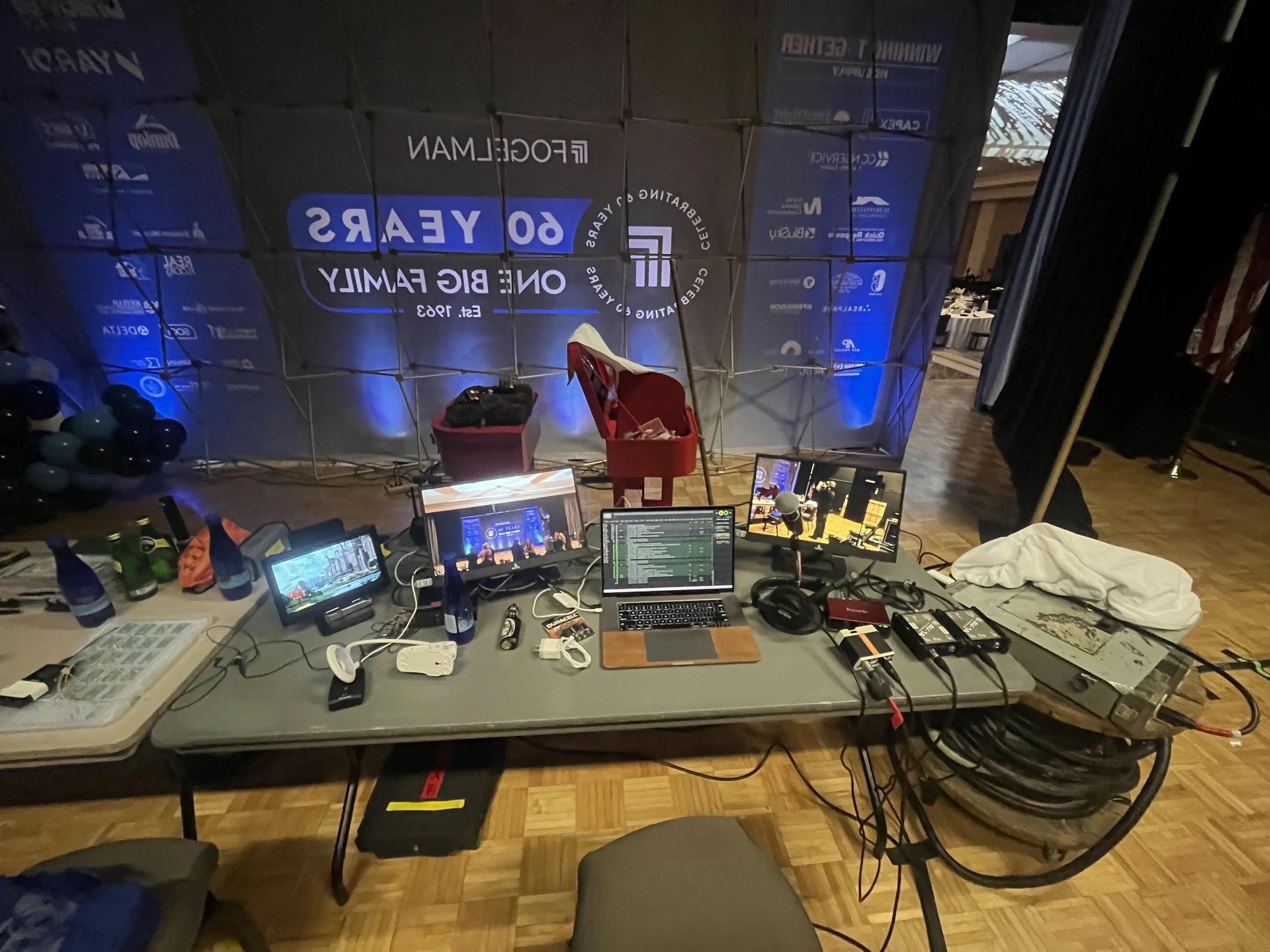 Event setup with multiple monitors, microphones, and equipment on a table in front of a banner that reads '60 Years, One Big Family, Est. 1963' at a venue with wooden floors.