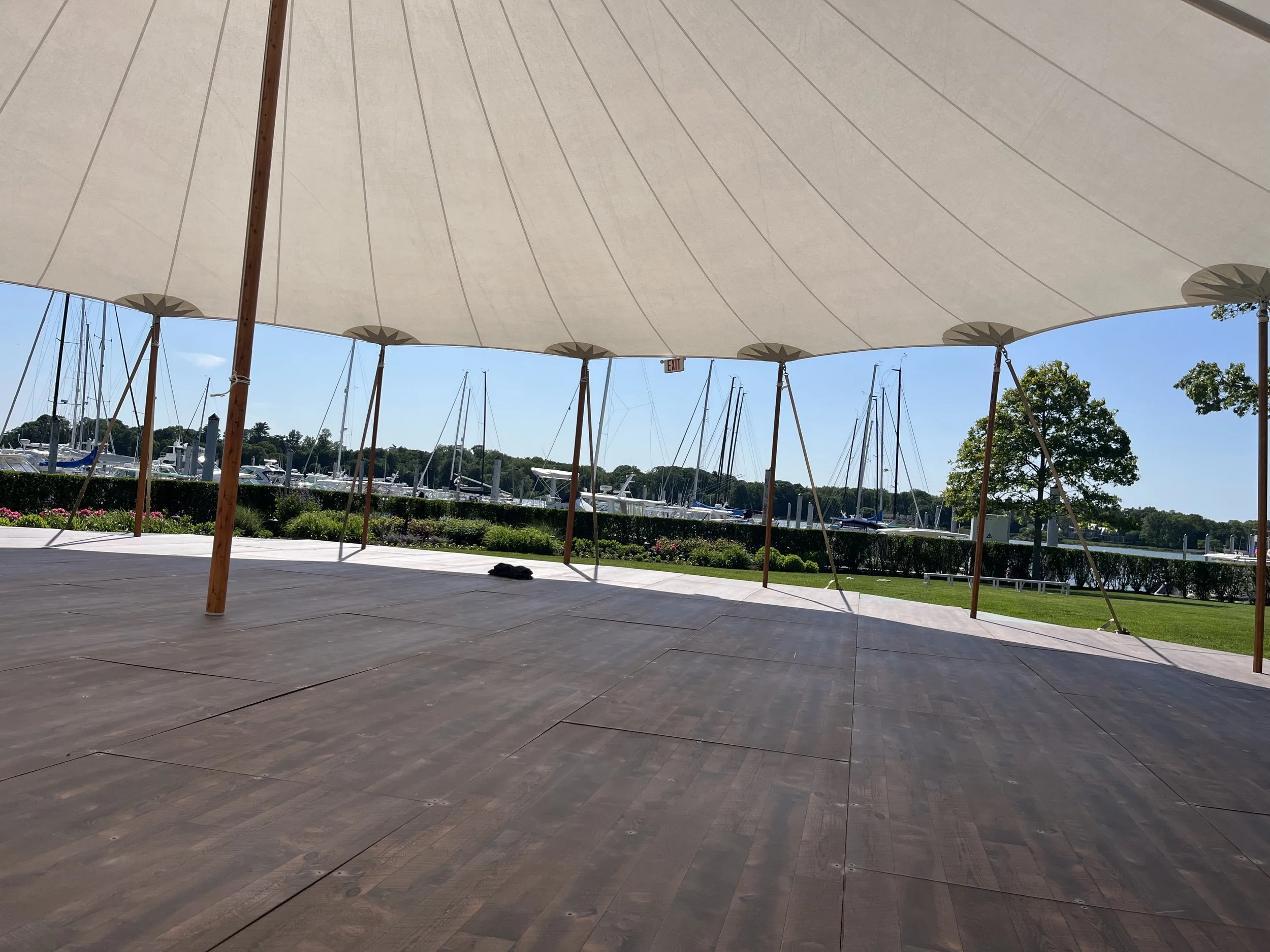 View of a marina with docked sailboats, taken from under a large white canopy with wooden supports, on a sunny day.