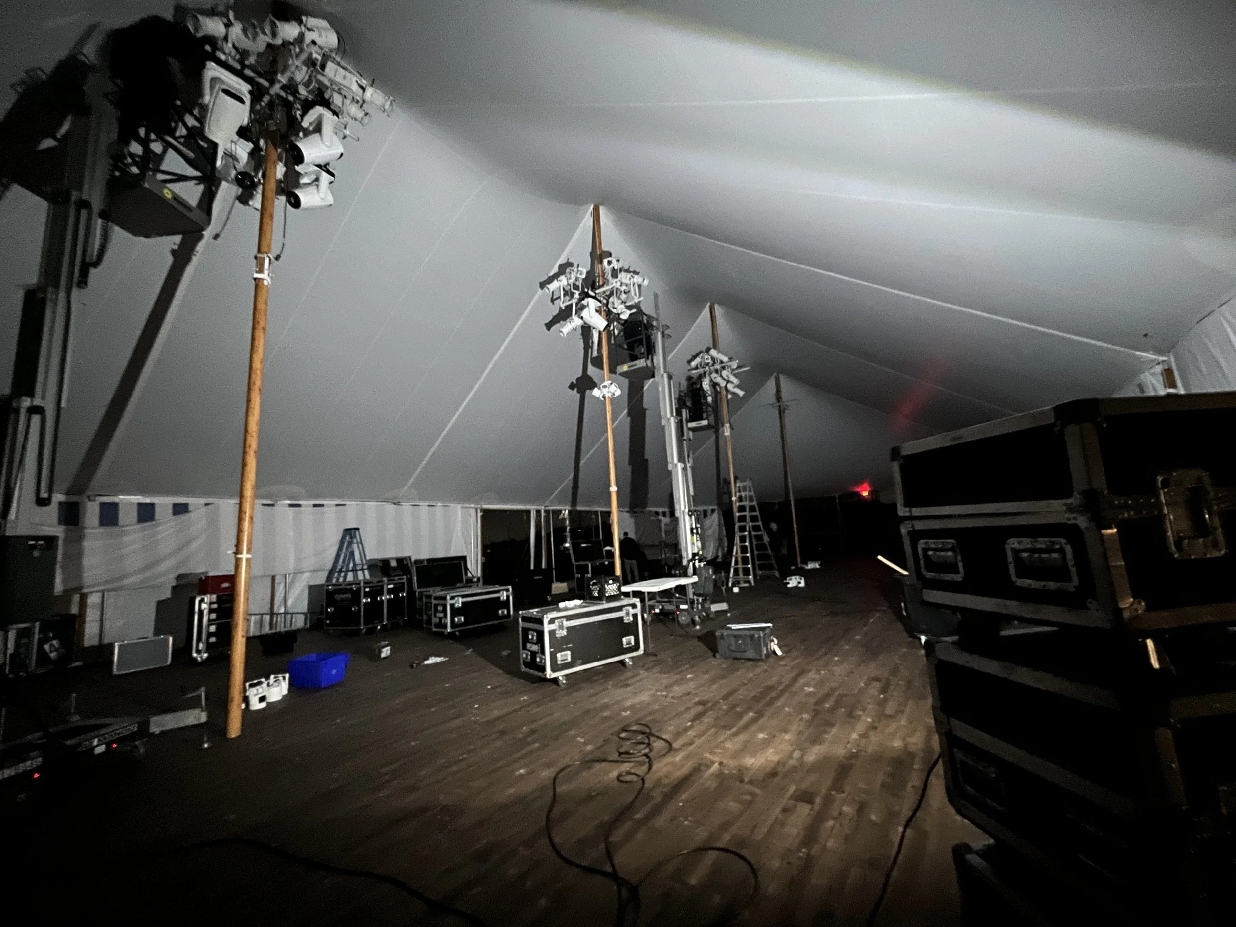 Stage lighting equipment, ladders, and storage boxes inside a large white tent, set up for an event or concert.