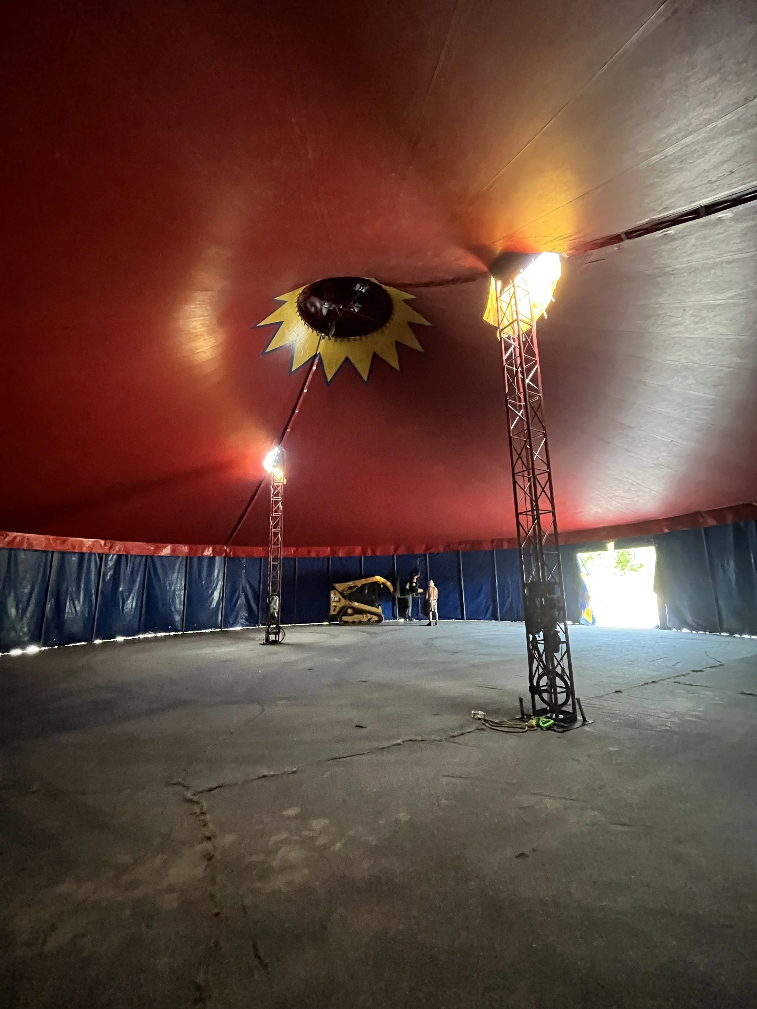 Interior of a circus tent with a red ceiling and a large opening that lets in natural light. There are two tall lighting poles and a small construction vehicle. A person is visible in the distance.