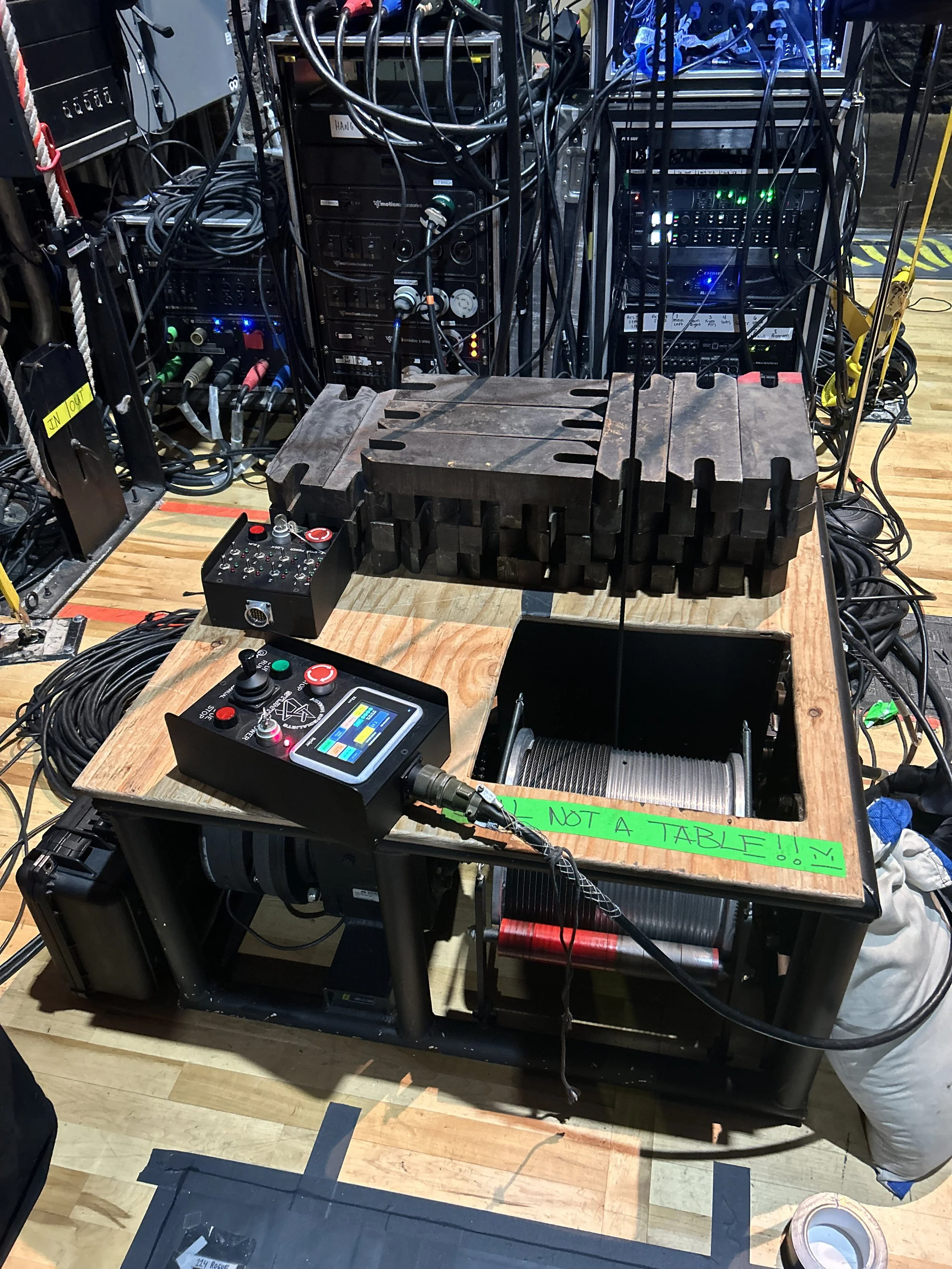 A wooden table with electronic equipment, cables, and foam blocks on top, surrounded by additional electronic devices and wires in a technical or sound setup. A bright green tape on the table reads "NOT A TABLE!!"