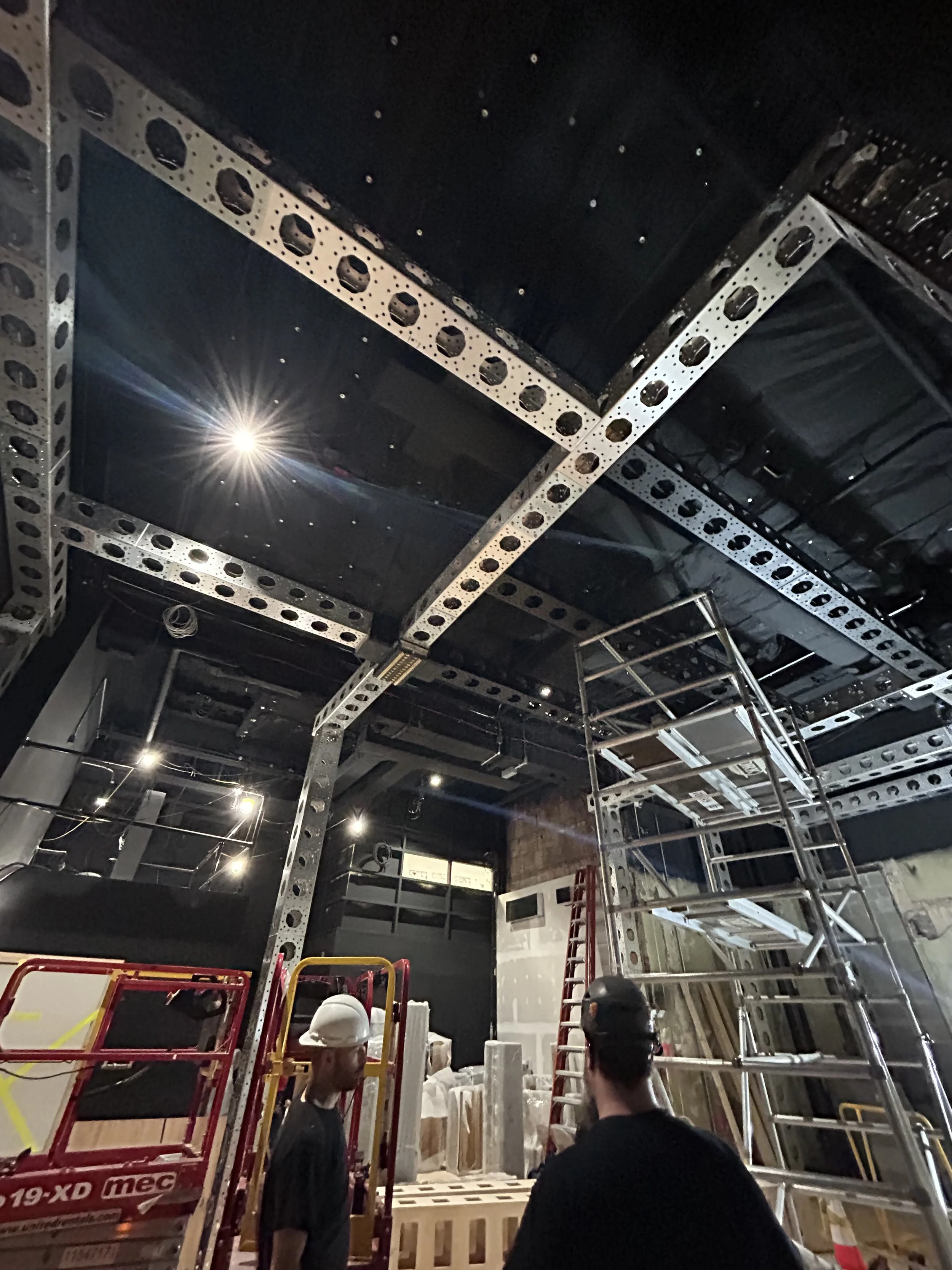 Construction workers in a building under renovation, with metal scaffolding, ladders, and construction materials visible, and a black ceiling with exposed beams and ductwork.