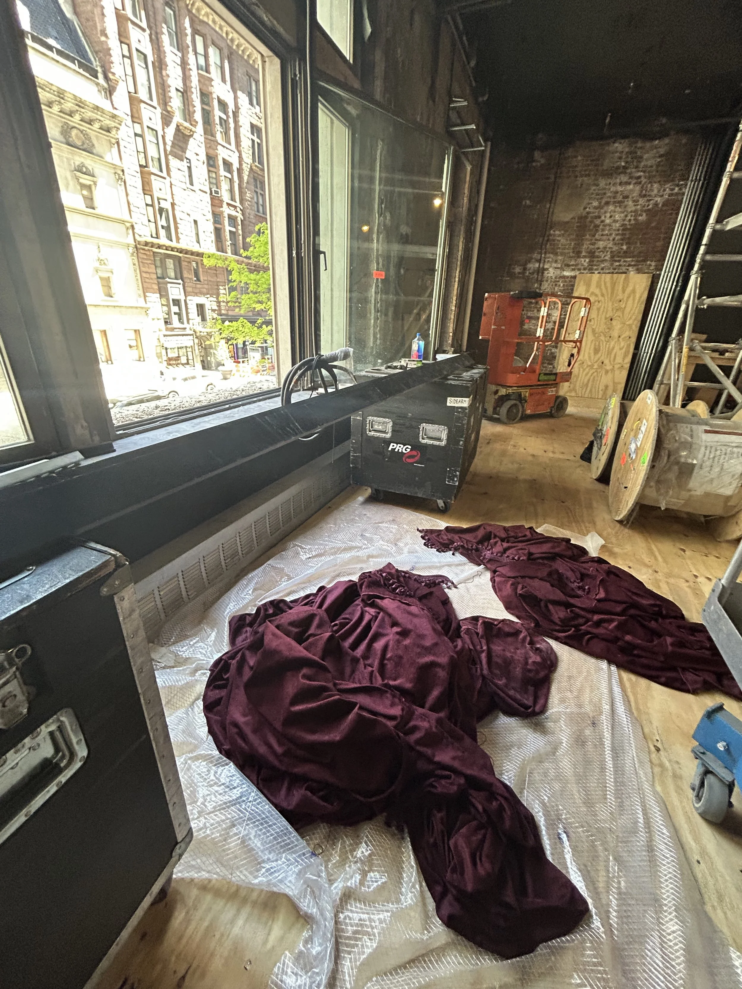 An interior view of a room under construction with large windows open to the street outside. The floor is covered with a protective sheet, and there are crumpled burgundy fabric and various construction materials, equipment, and tools scattered aroun