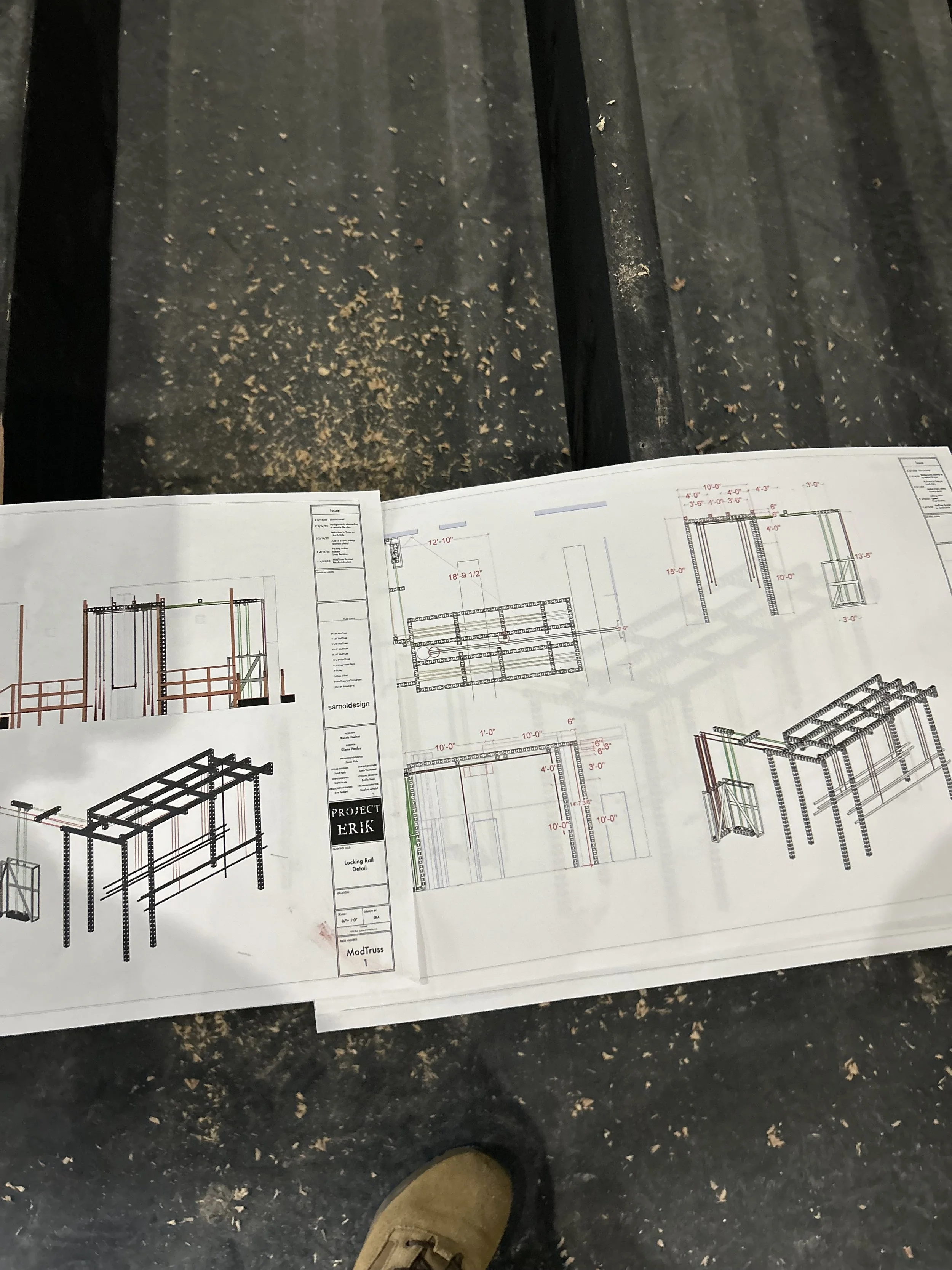 Construction blueprint or technical drawing on paper showing structural details, resting on a black surface with wood shavings. A person's tan boot is visible at the bottom of the image.