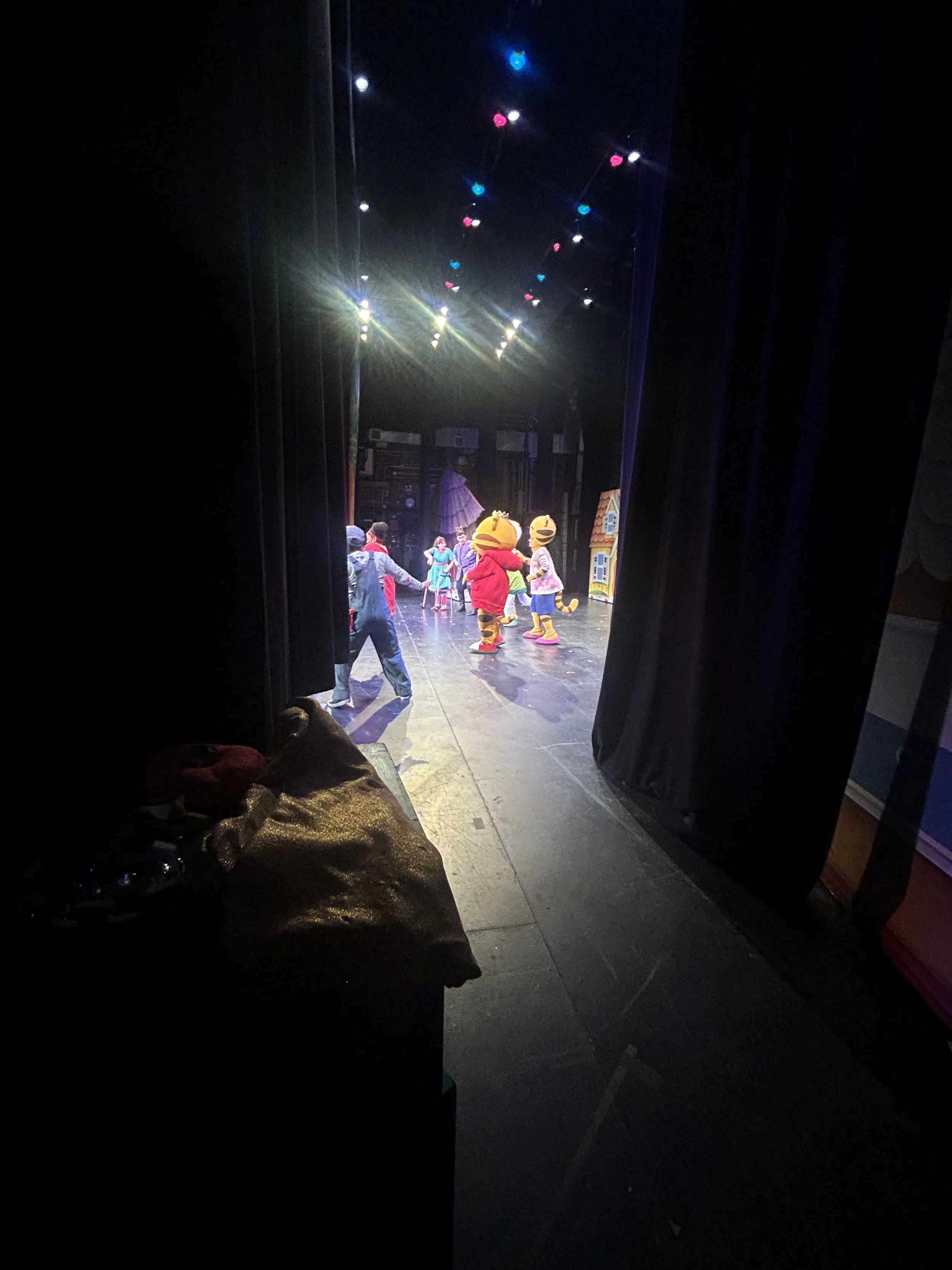 Children performing on a stage with characters in costumes, seen through an opening in the curtain, stage lighting overhead.