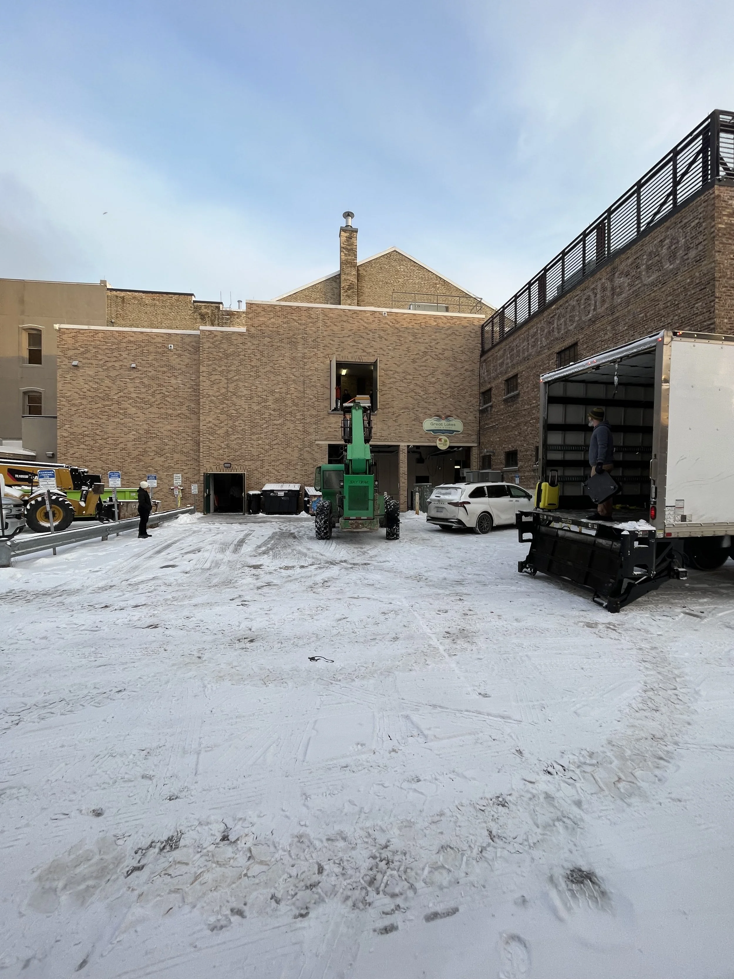 Snow-covered parking lot with a green lift equipment, a white SUV, a white truck with an open side door, and a man standing inside the truck. Buildings with brick facades are in the background under a partly cloudy sky.
