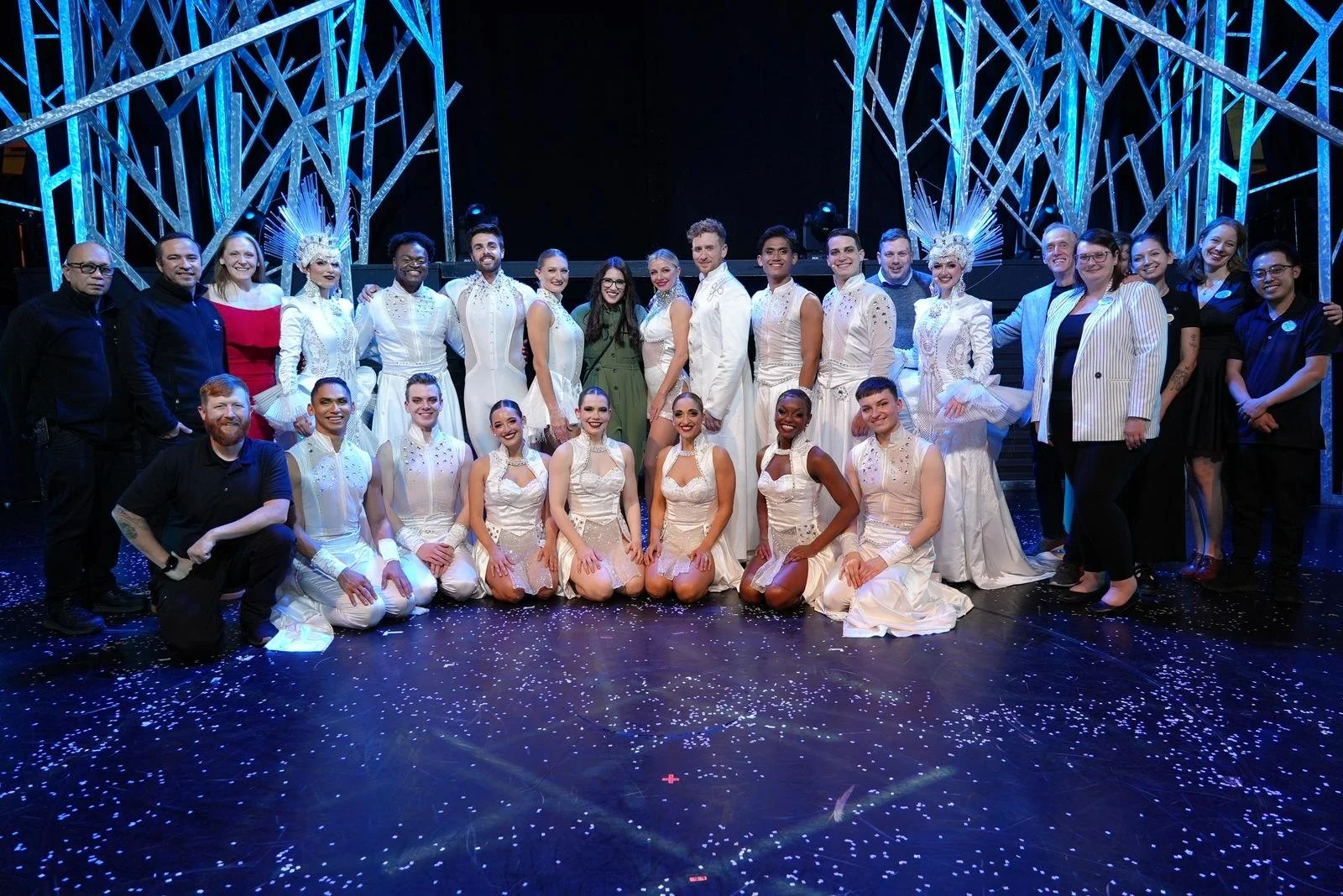 A large group of performers and crew members posing on stage after a show, with some wearing elaborate white costumes and headpieces, and others in casual or team attire, under blue stage lighting with a decorative geometric backdrop.