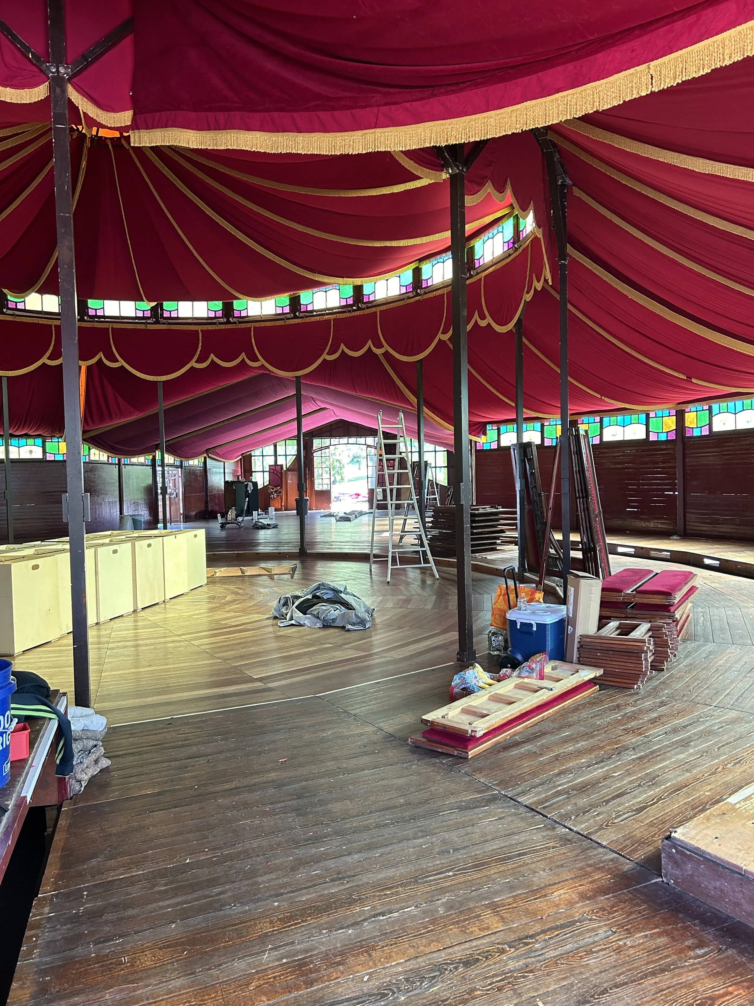 Inside a decorated tent with red and gold drapes, construction supplies and a ladder, indicating ongoing setup or renovation.
