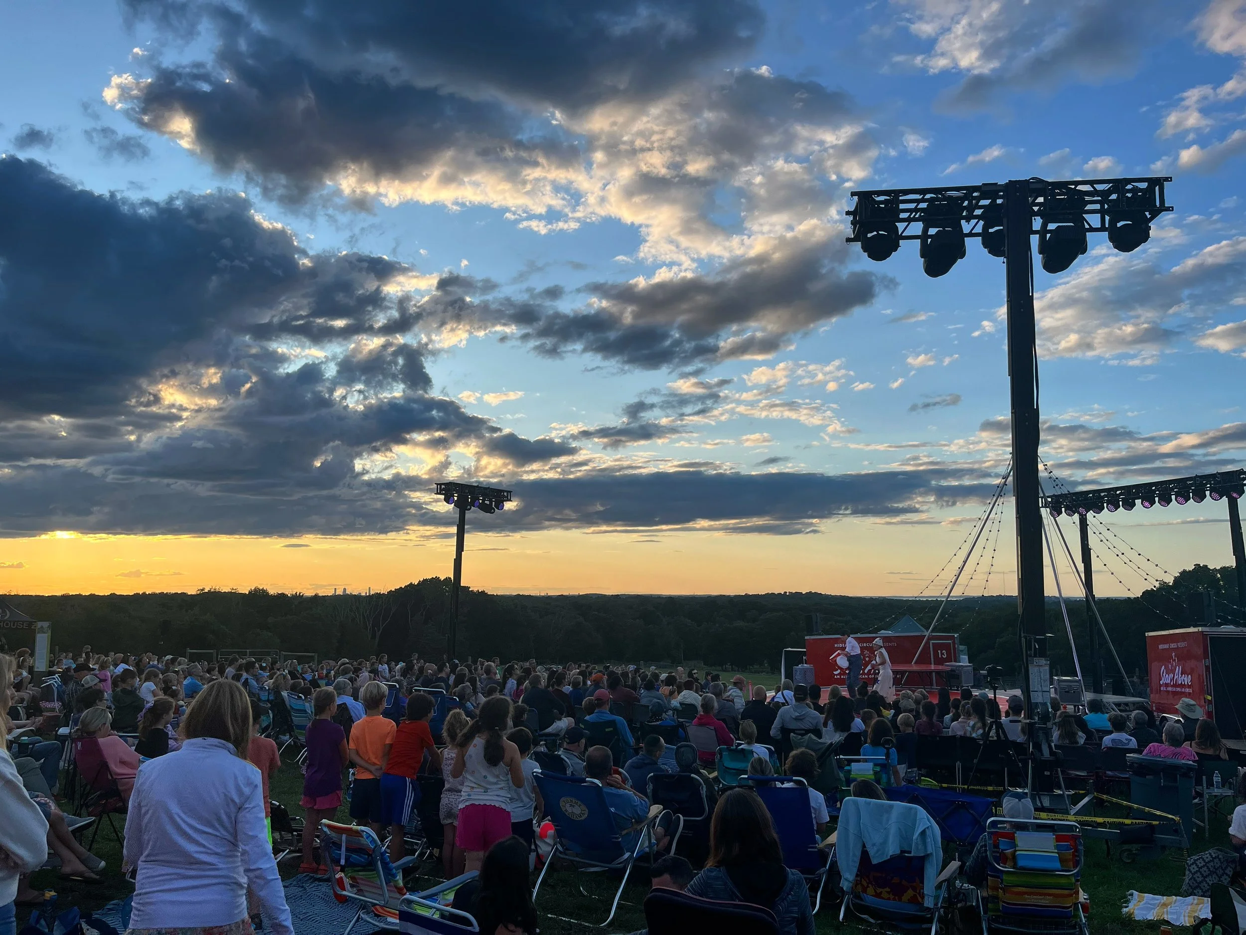 An outdoor event at sunset with a large audience seated on chairs and blankets, watching a stage with performers. The sky has dramatic clouds and the sun is setting on the horizon.