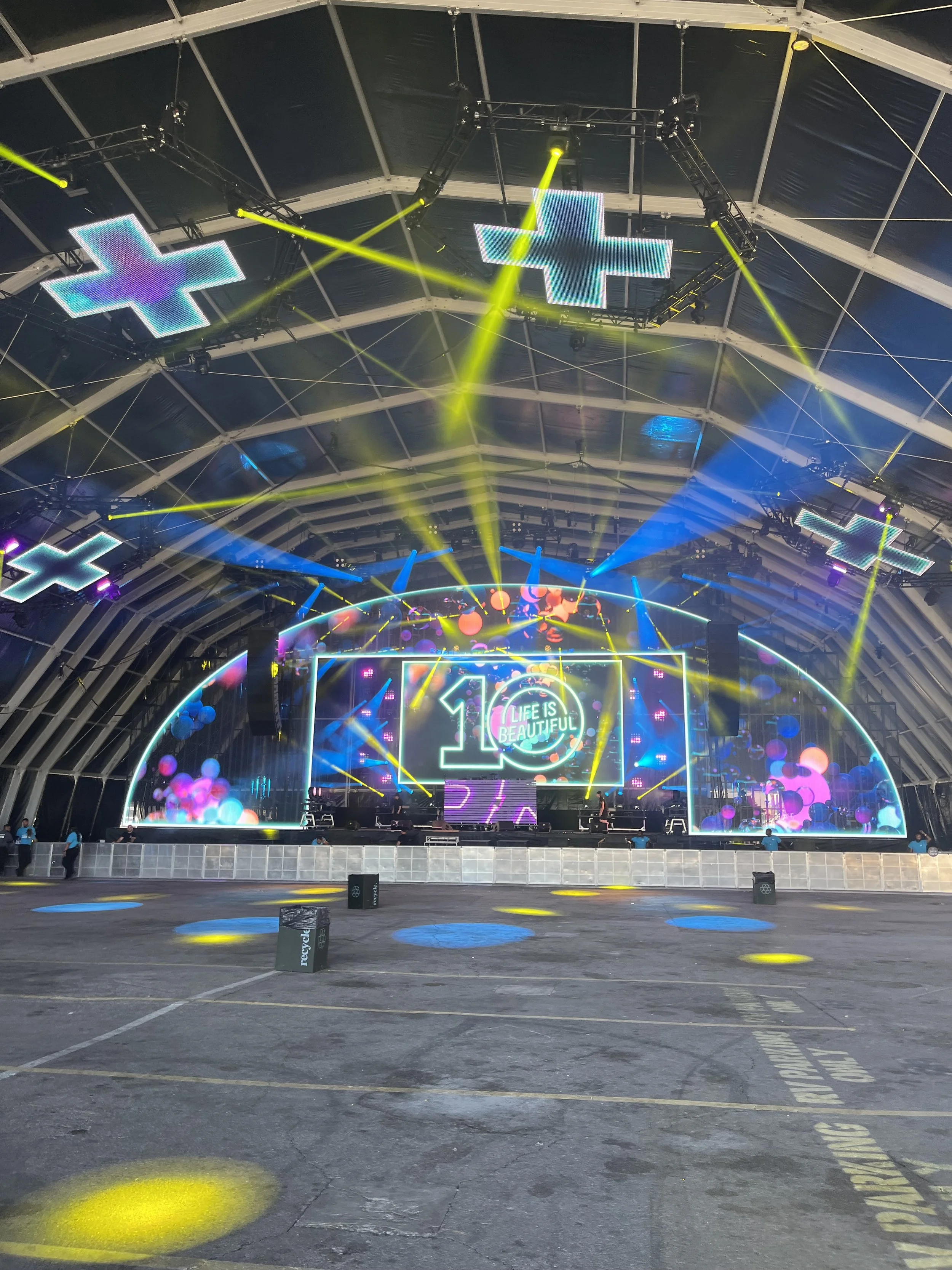 Empty stage with colorful LED screens displaying '10 Life is Beautiful' at a large indoor concert venue, with yellow and blue stage lights and fog effects.
