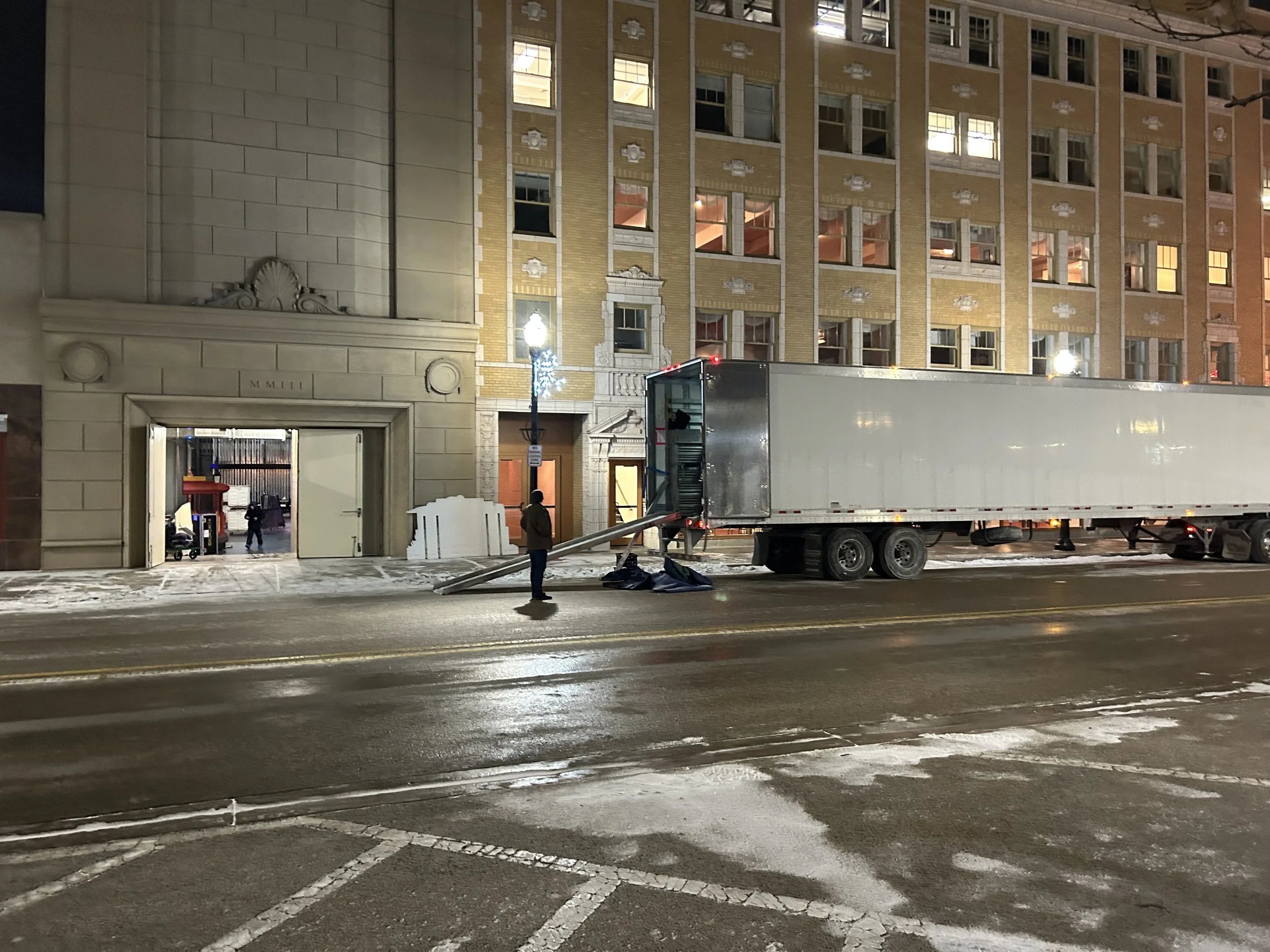 Nighttime image of a large white truck parked on a city street in front of a multi-story brick building. The truck’s trailer door is slightly open, with a long metal ramp extending to the sidewalk. A person stands near the front of the truck, and the