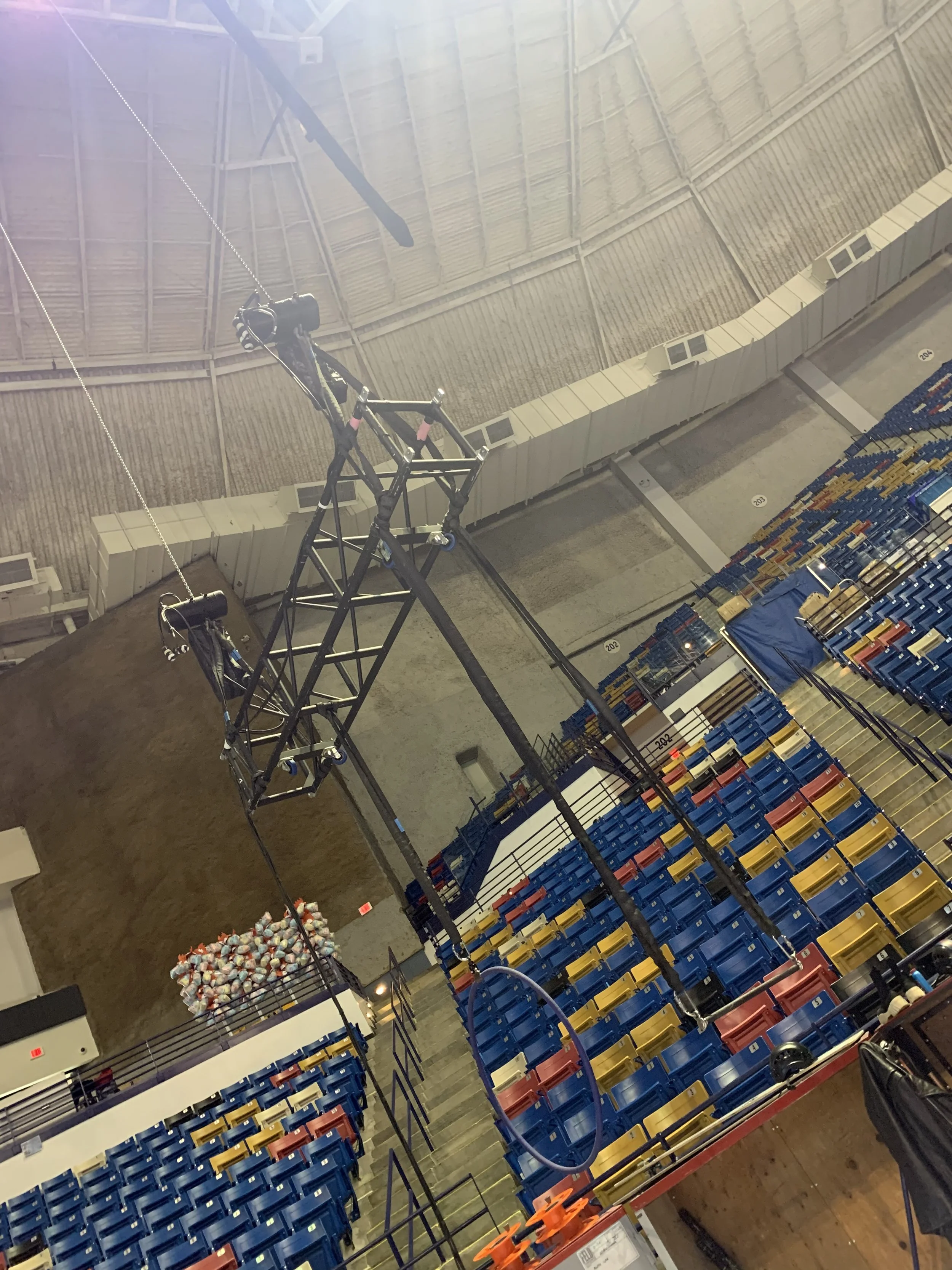 An indoor arena with empty blue, yellow, and red seats. A large black metal structure is suspended from the ceiling. There are basketball hoops and a large mound of what appears to be dirt or clay at the back, with a pile of small objects, possibly r