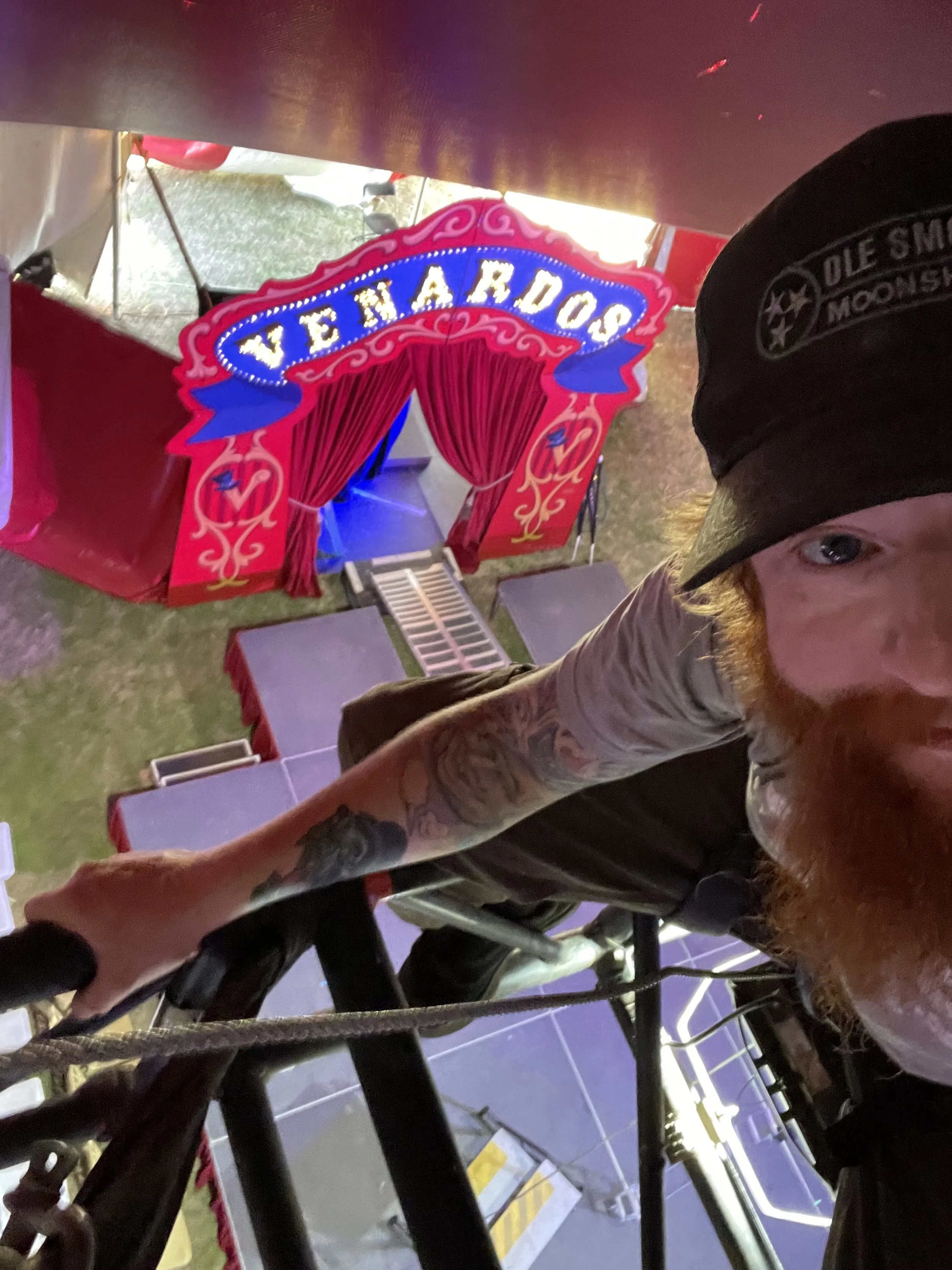 A man with a red beard and tattoos takes a selfie from a ladder or elevated position, with a colorful circus or carnival sign in the background that reads 'VENA RDOC' with red curtains and decorative elements underneath.
