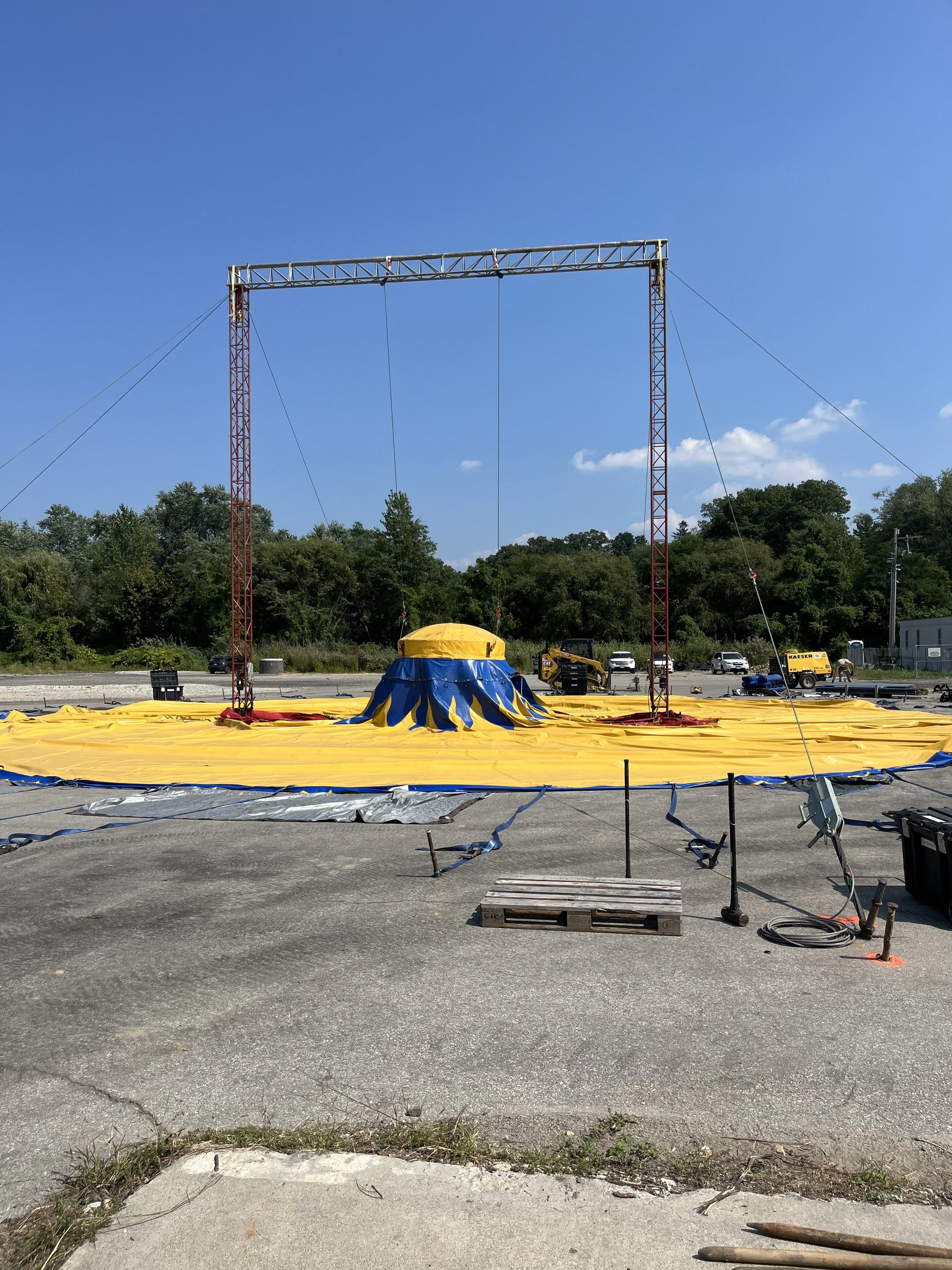 An amusement park ride, likely a carnival or circus attraction, being assembled outdoors on a paved area. The ride has a central yellow and blue canopy with a large, flat, circular yellow fabric surrounding it, secured to a red metal frame structure