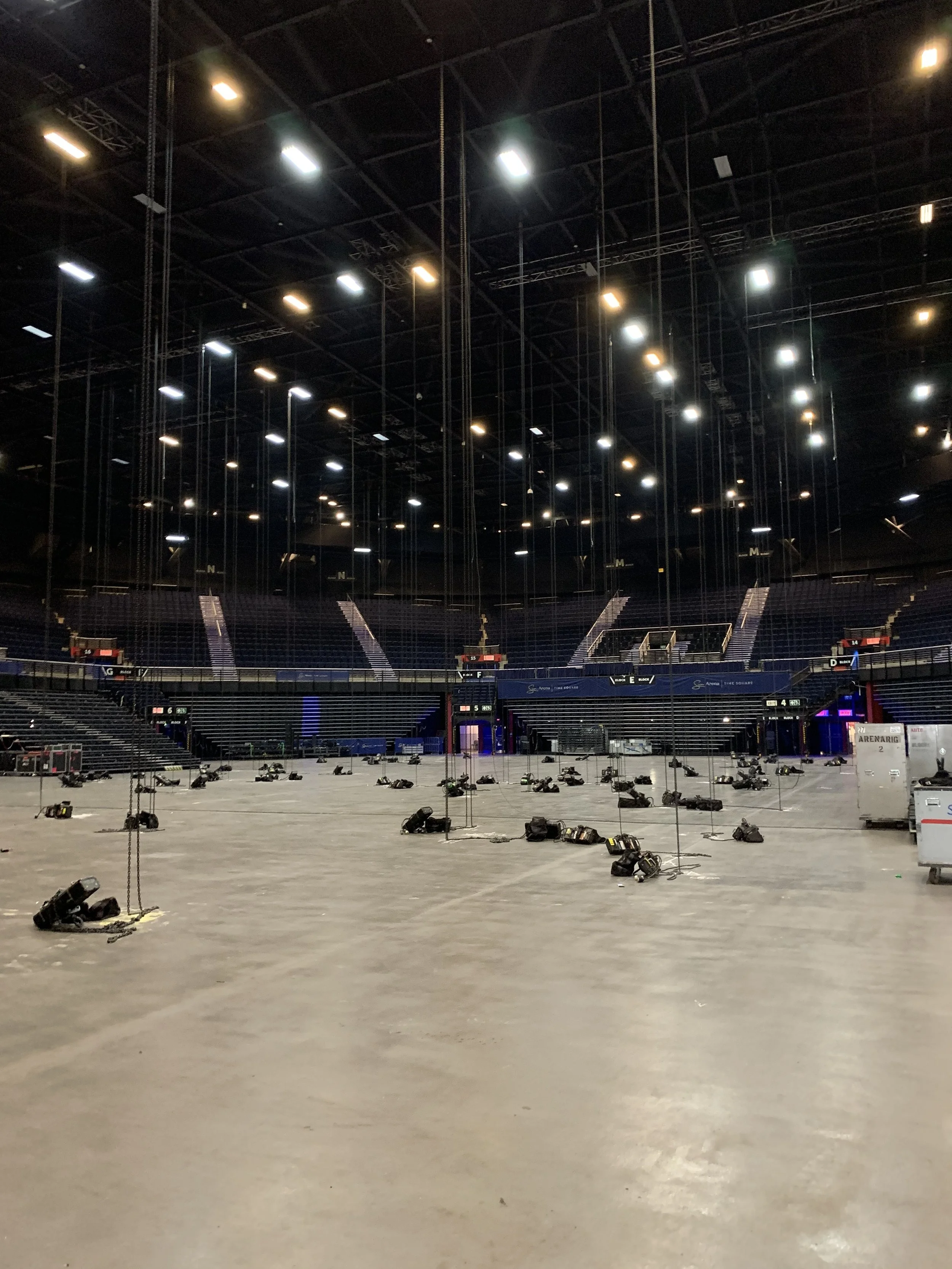 Inside an empty sports arena with stage lighting and seating, scattered with equipment and lighting fixtures on the floor.