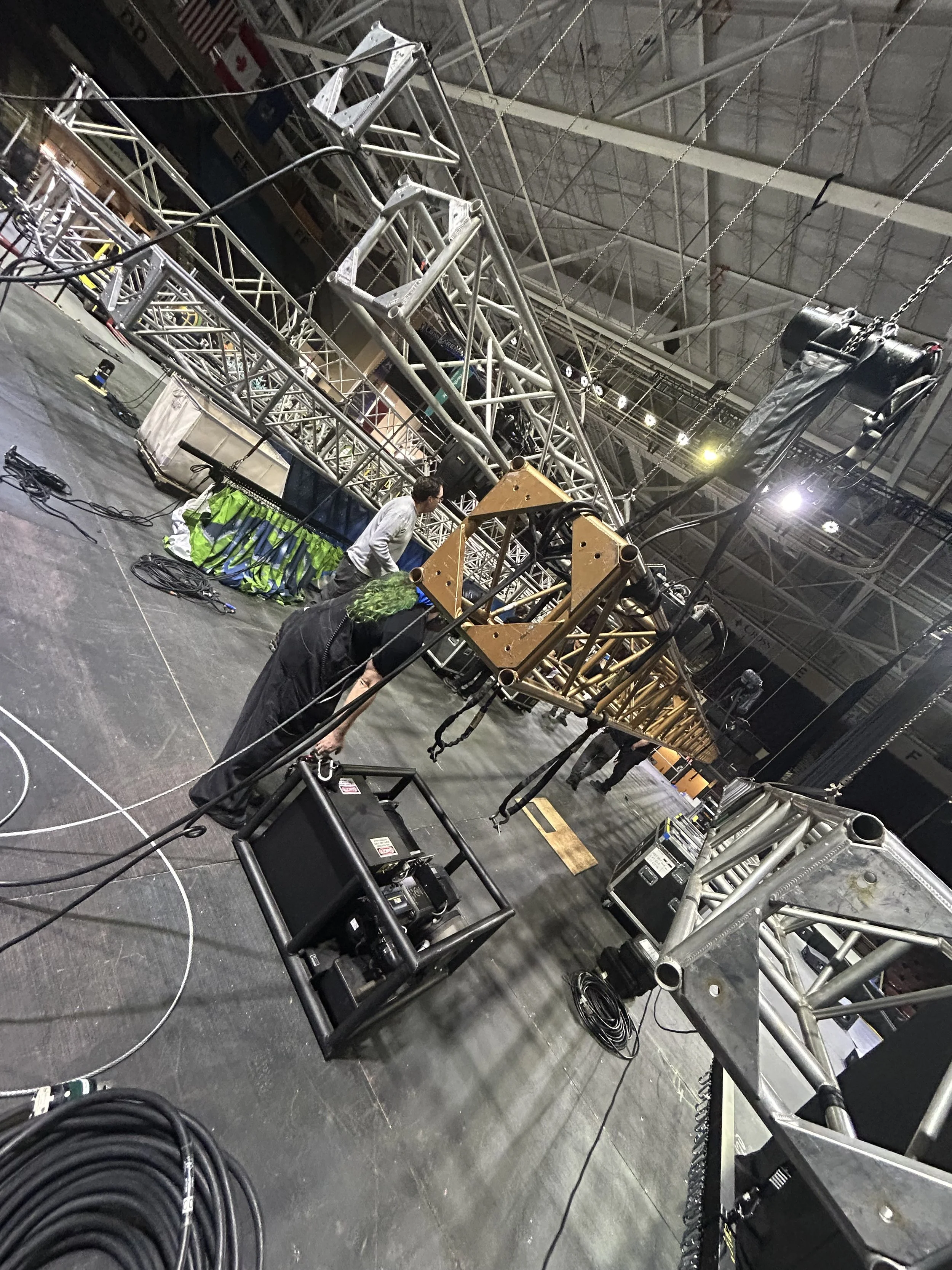 Workers assembling lighting and truss structures on a stage in a large indoor venue.