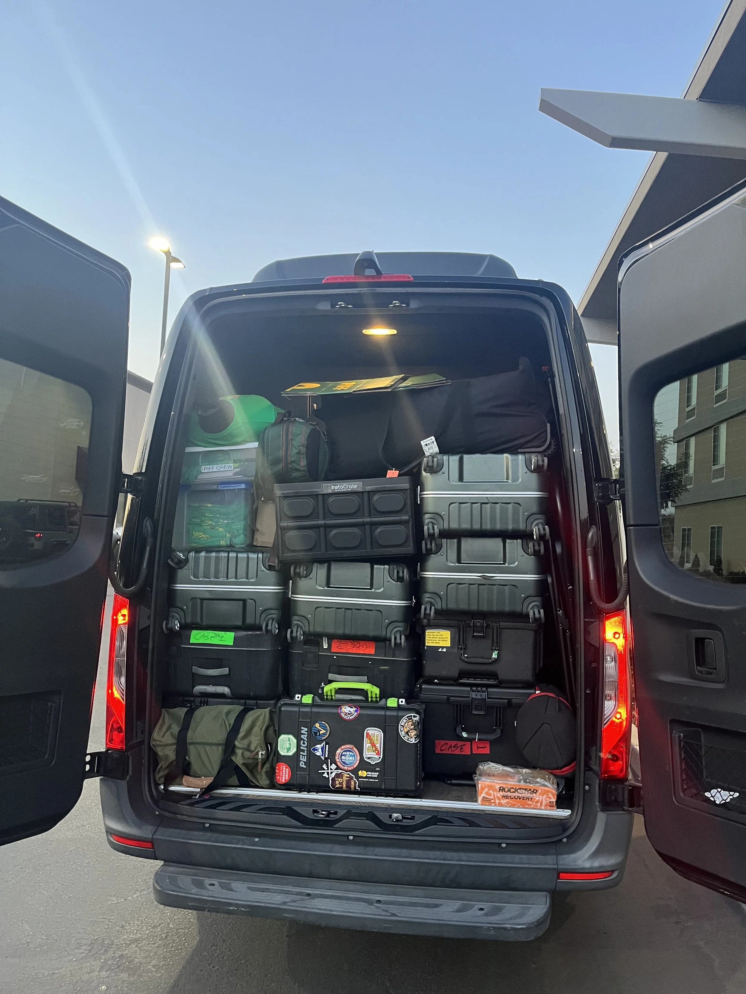 Open rear of a vehicle filled with various suitcases, bags, and gear, parked outside with building and sky in background.