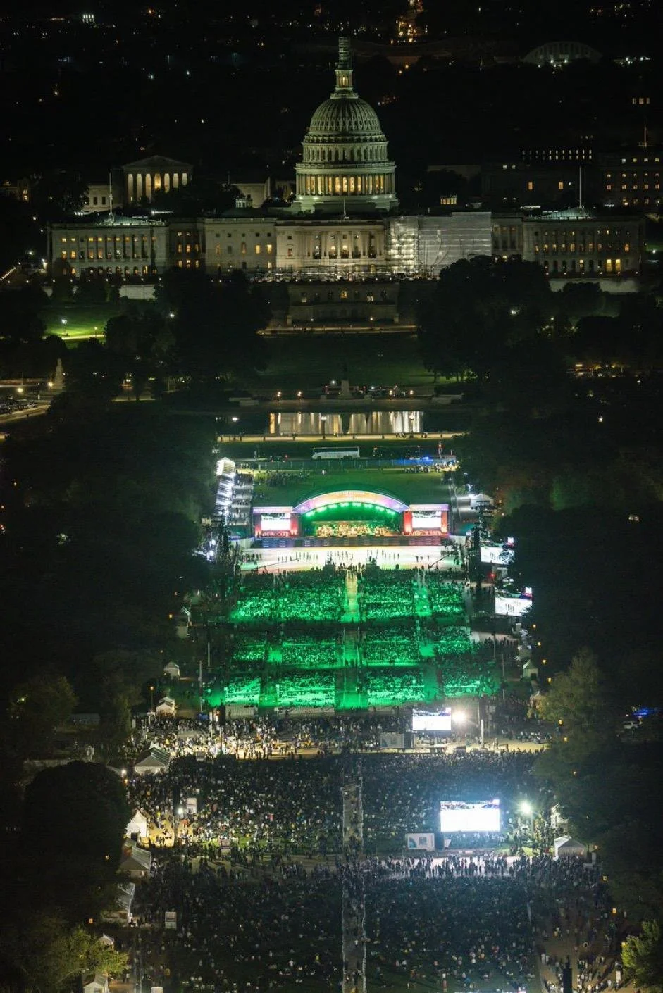 Nighttime view of the U.S. Capitol building illuminated, with a large crowd gathered at the National Mall, featuring a stage with bright lights and green lighting effects.