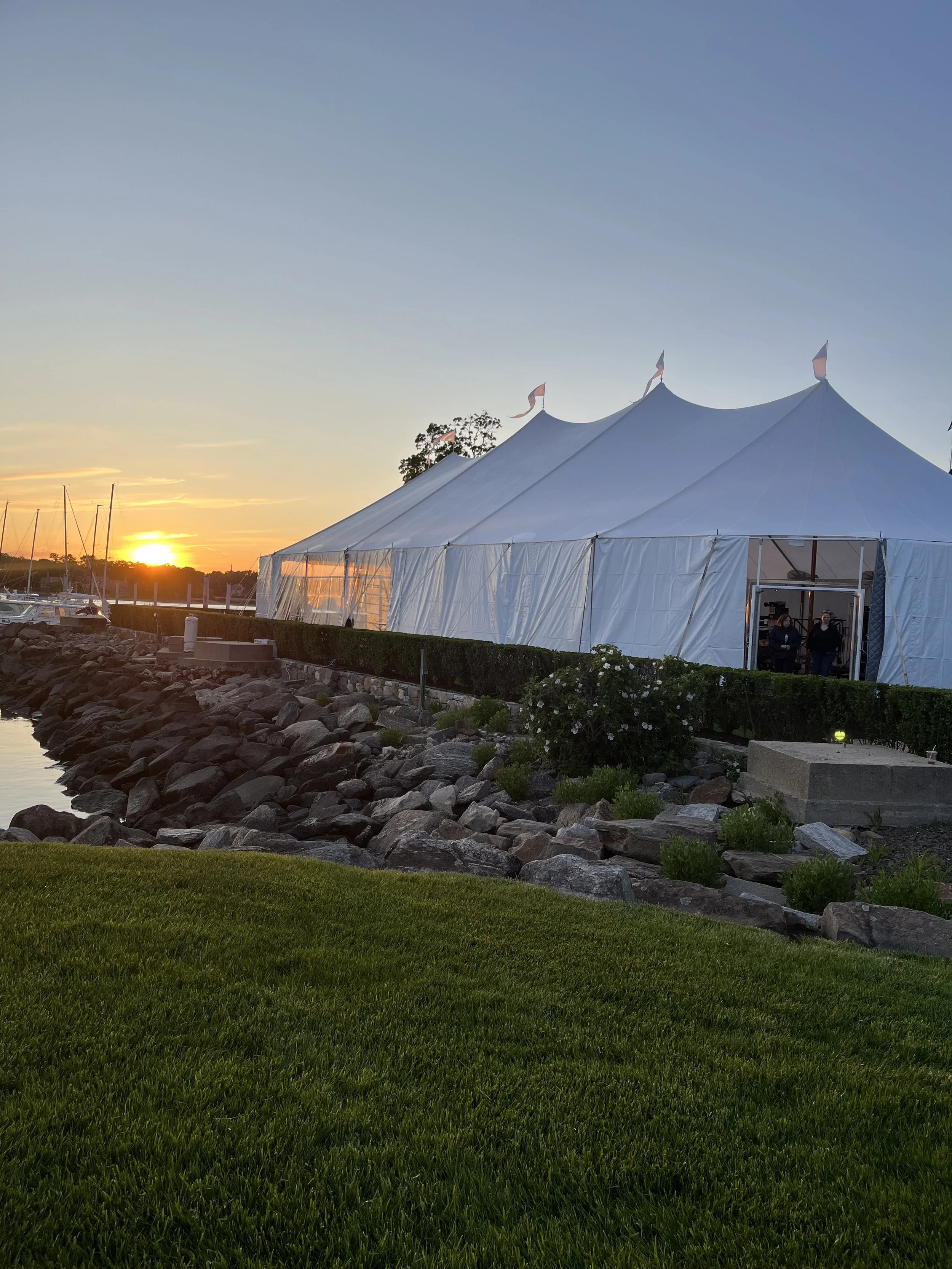 A large white event tent set up near a body of water, with sailboats docked nearby, during sunset.