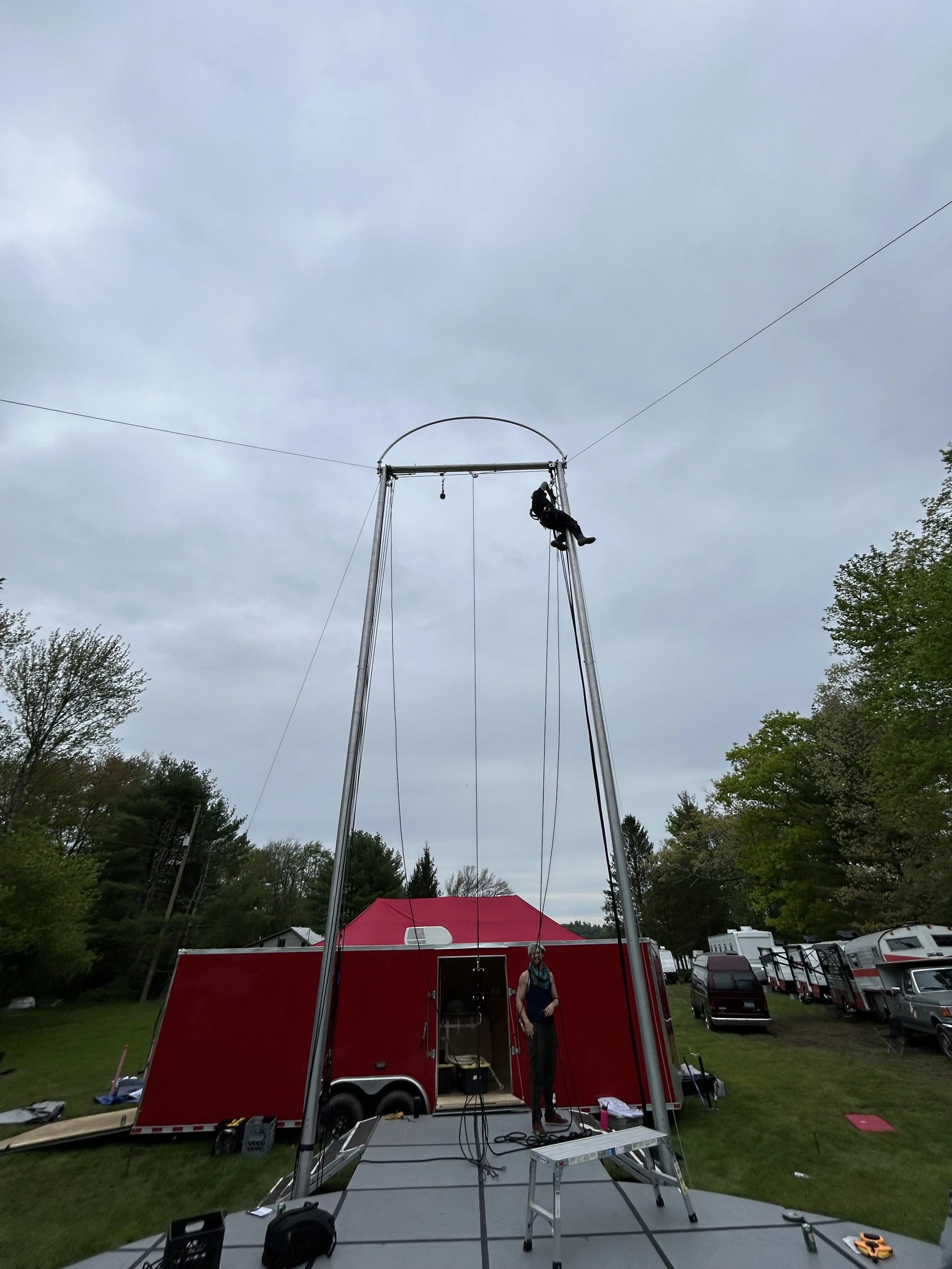 A person climbing a tall metal structure for a bungee jump setup, with a red trailer and trees in the background on a cloudy day.