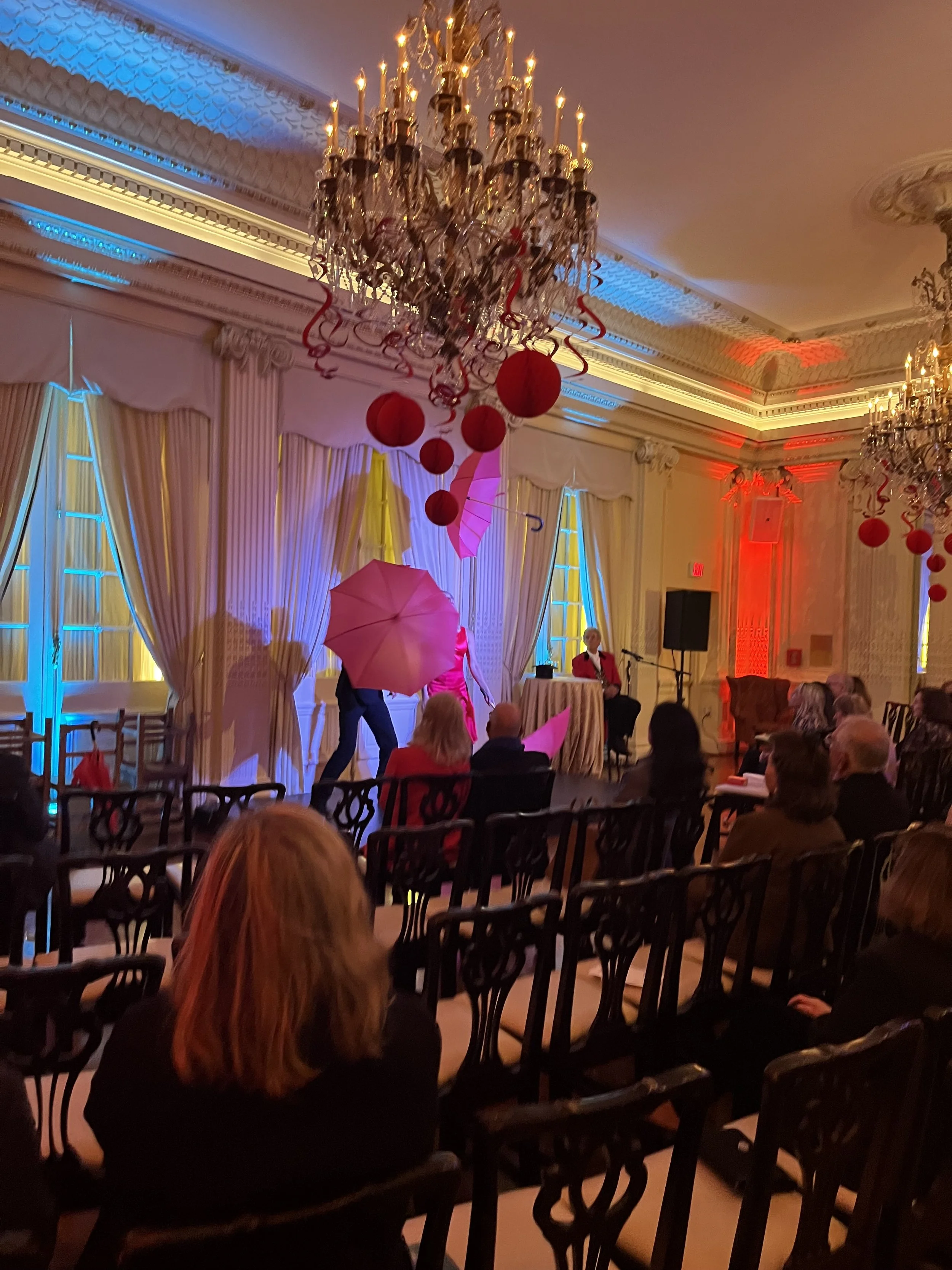 A decorated event hall with chandeliers, red and pink umbrellas, and red hanging paper lanterns. People are seated watching a performance on stage.
