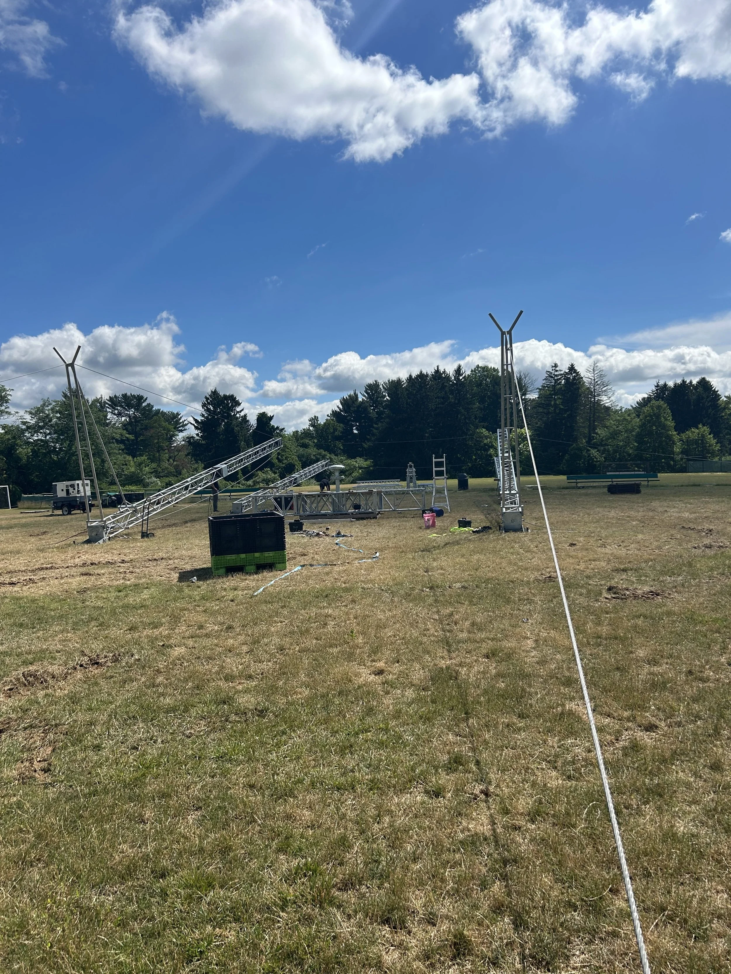 Disassembled metal tower structure on a grassy field, with equipment and a utility vehicle, under a blue sky with scattered clouds.