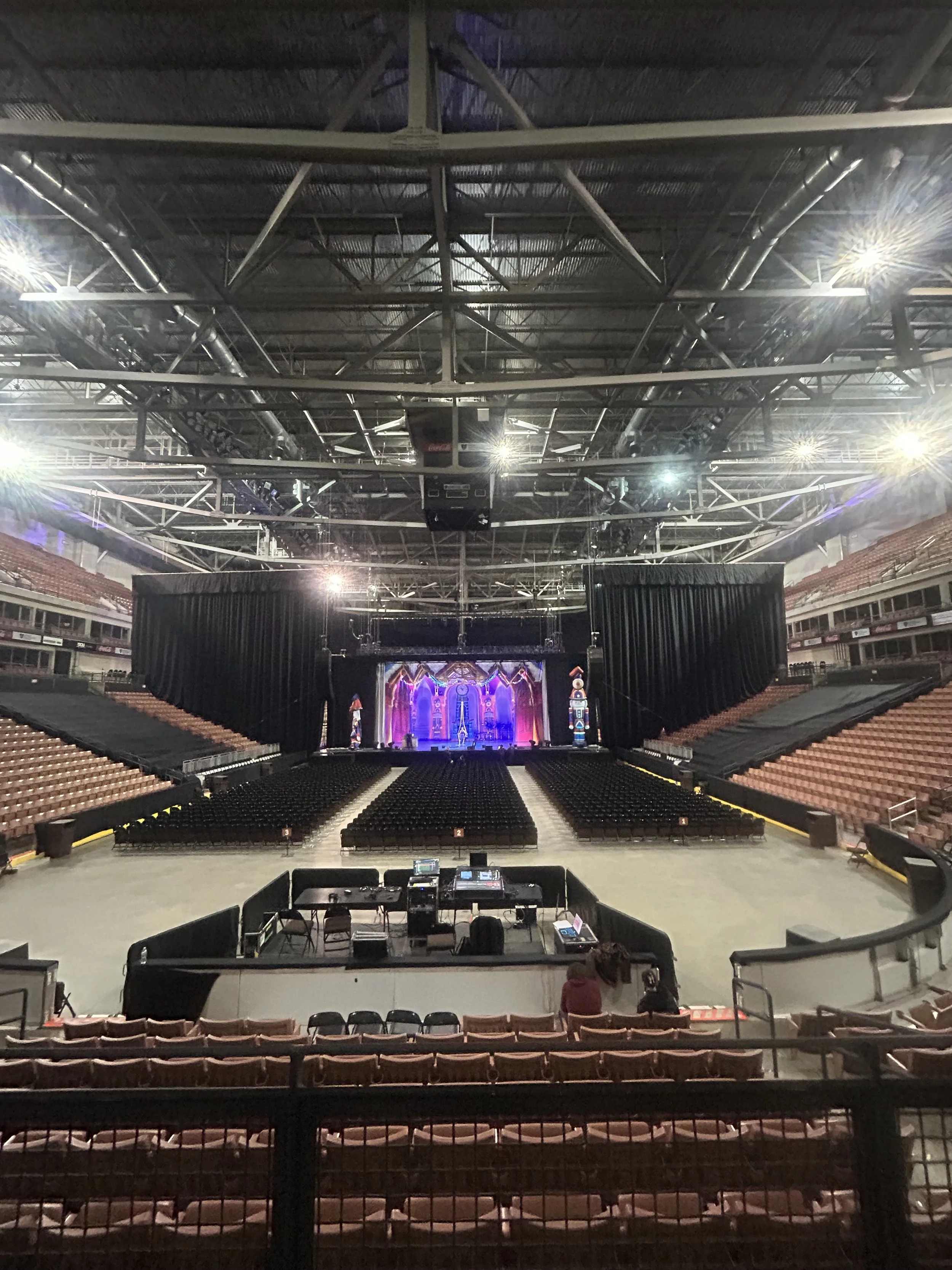 Inside a large arena or concert hall with empty seats, a stage with colorful lighting, and sound and lighting equipment set up in front.