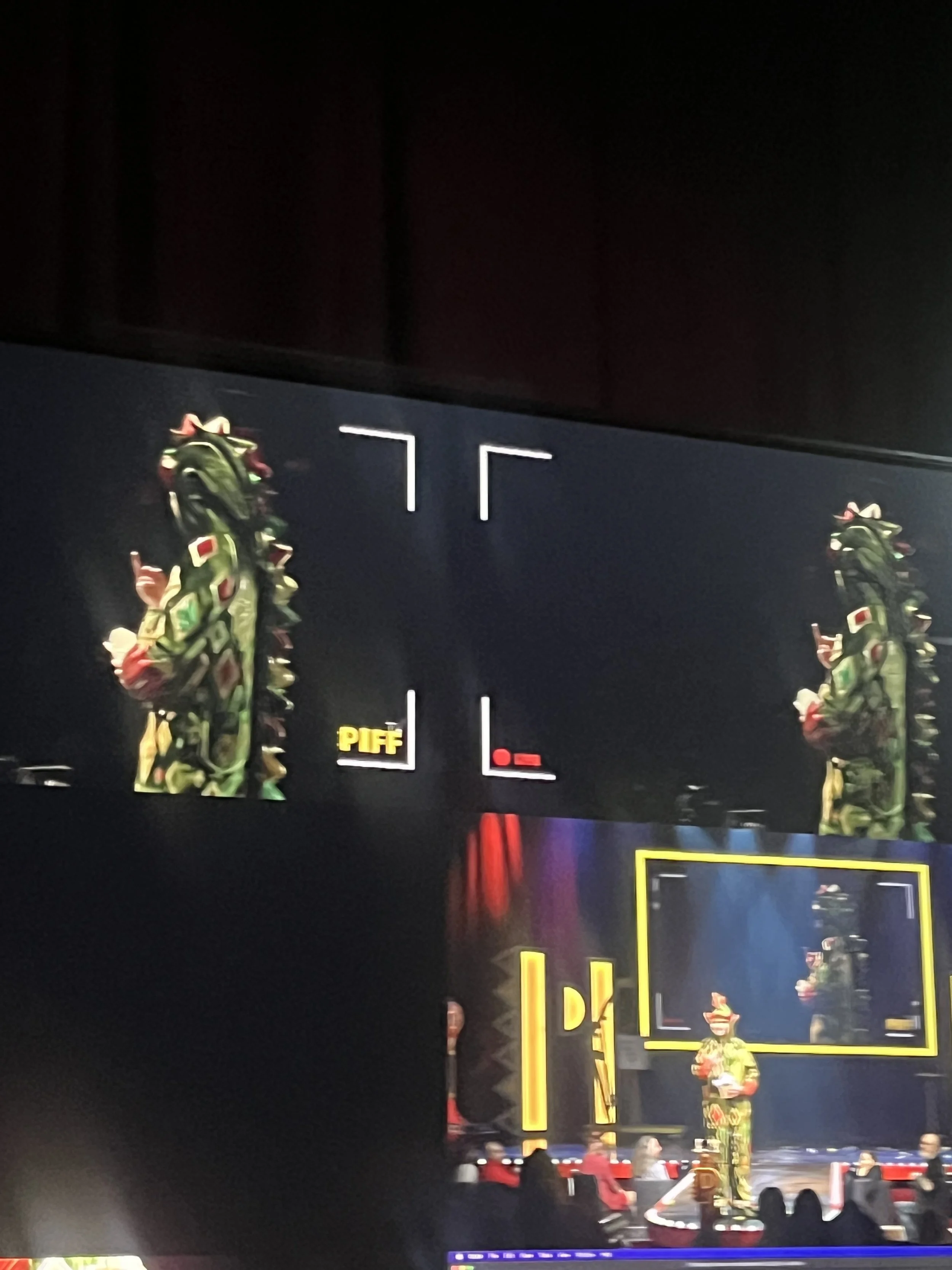 Stage with performer dressed in dinosaur costume, with large TV screens displaying the performer, and yellow decorative lights.