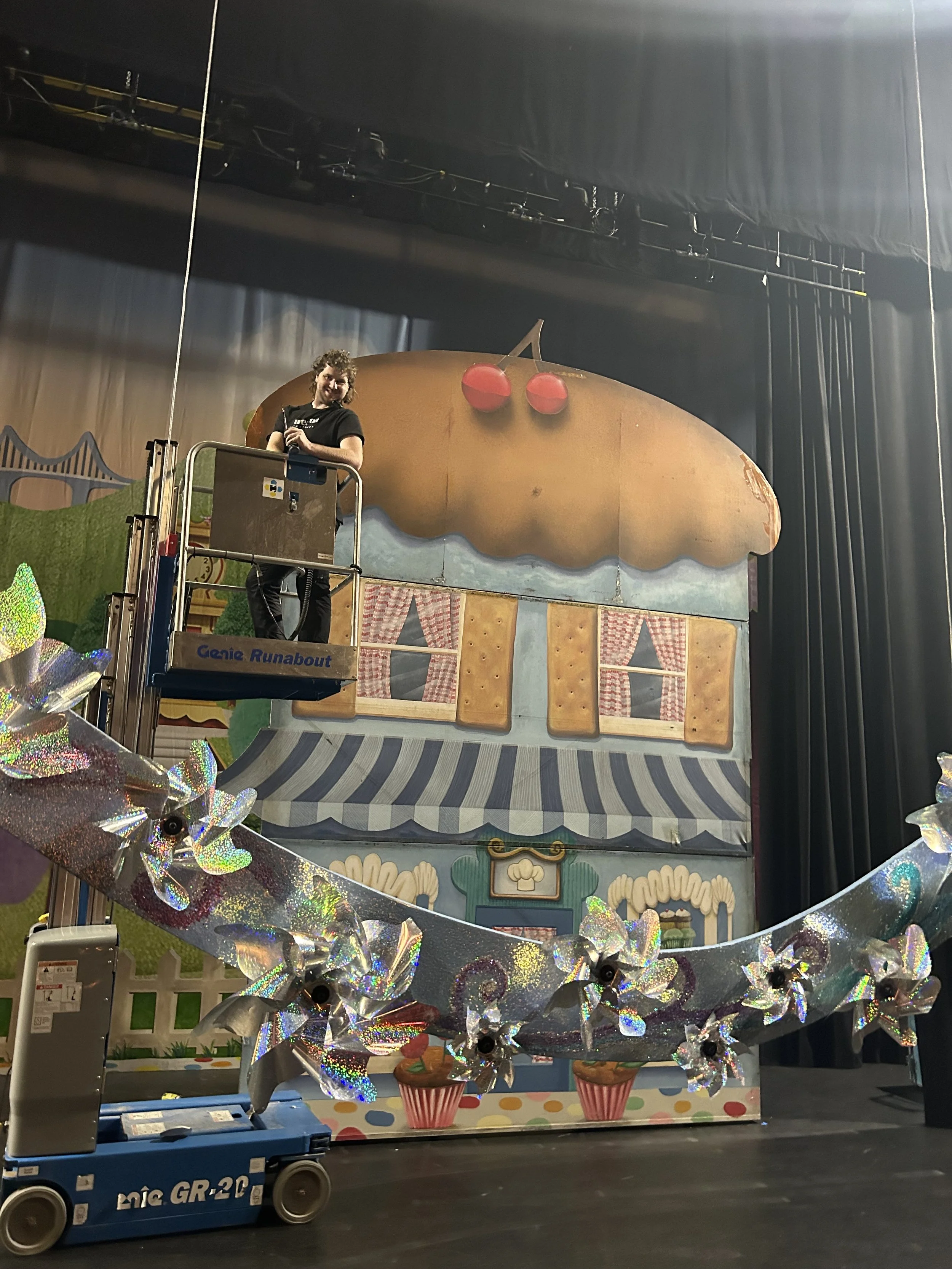Stage set with house facade resembling a cupcake with cherries on top, decorated with curtains and frosting details, and a person on a lift cleaning or inspecting the setup.