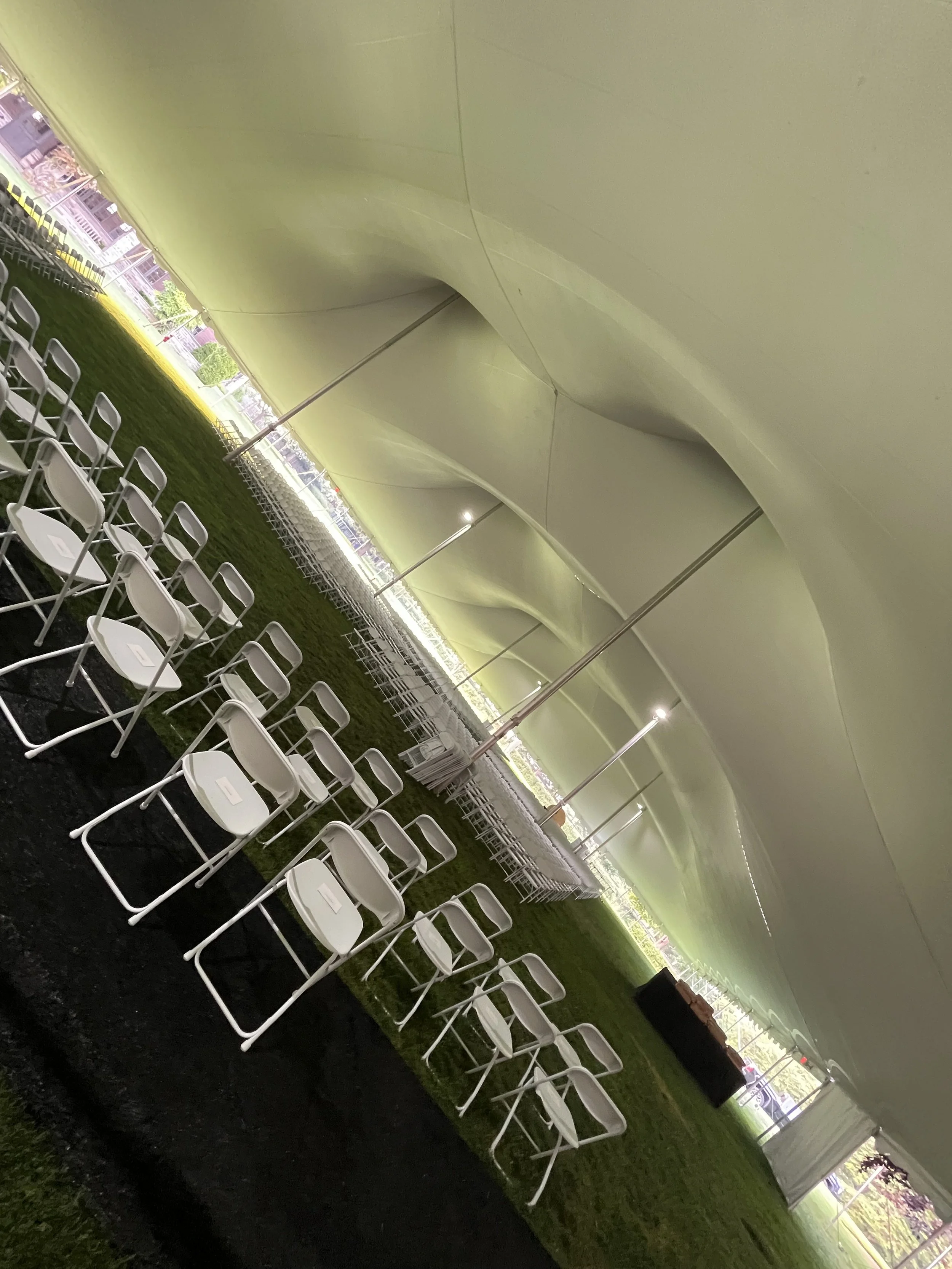 Empty white chairs arranged under a large tent on grass, with stage equipment in the background.
