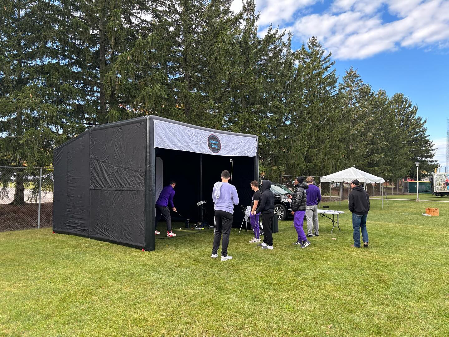 Group of people practicing golf on a small outdoor driving range with a black tent and trees in the background.