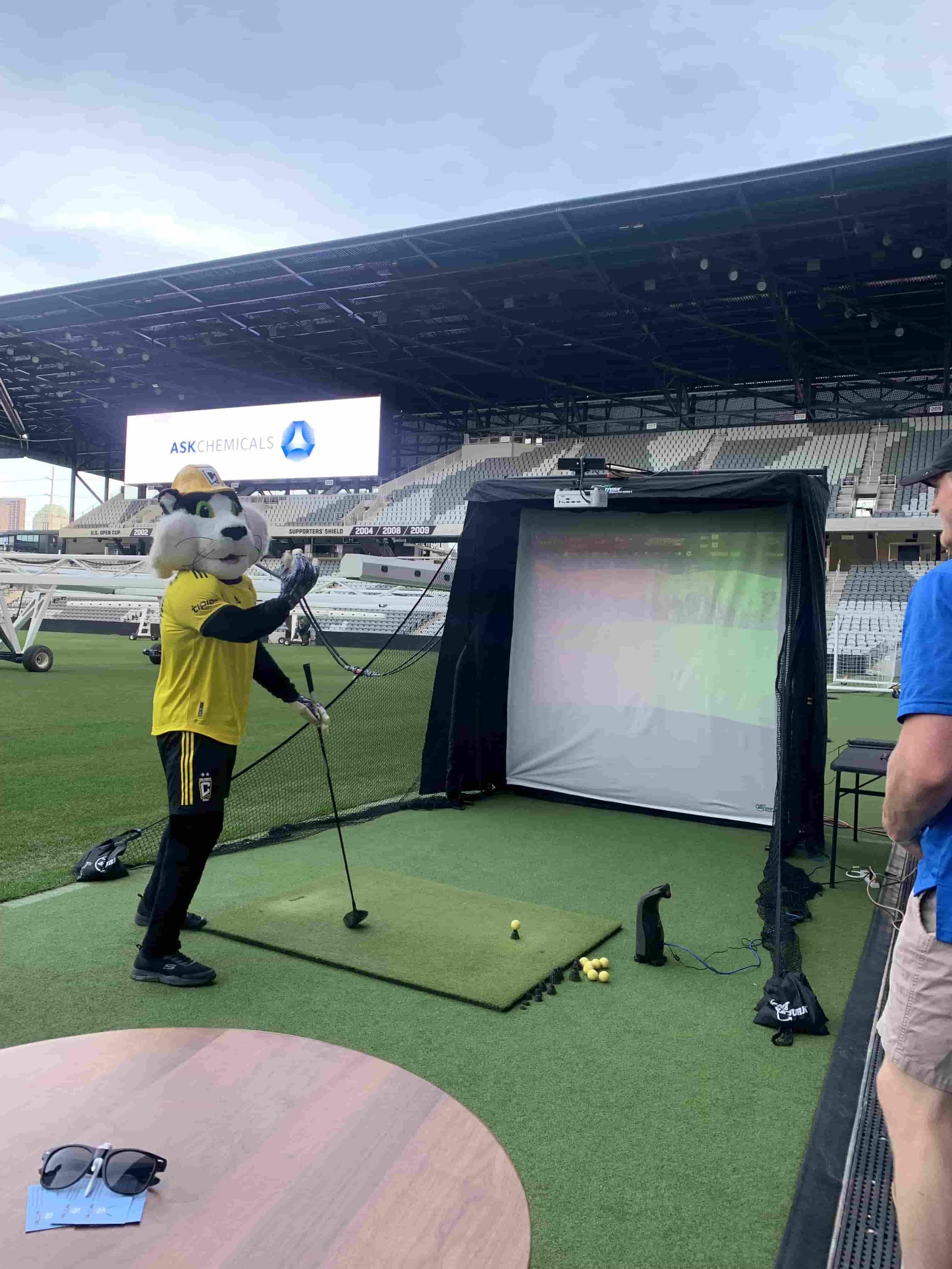 A person in a mascot costume practicing golf at a golf practice area, with a large screen displaying "ASK CHEMICALS" in the background.