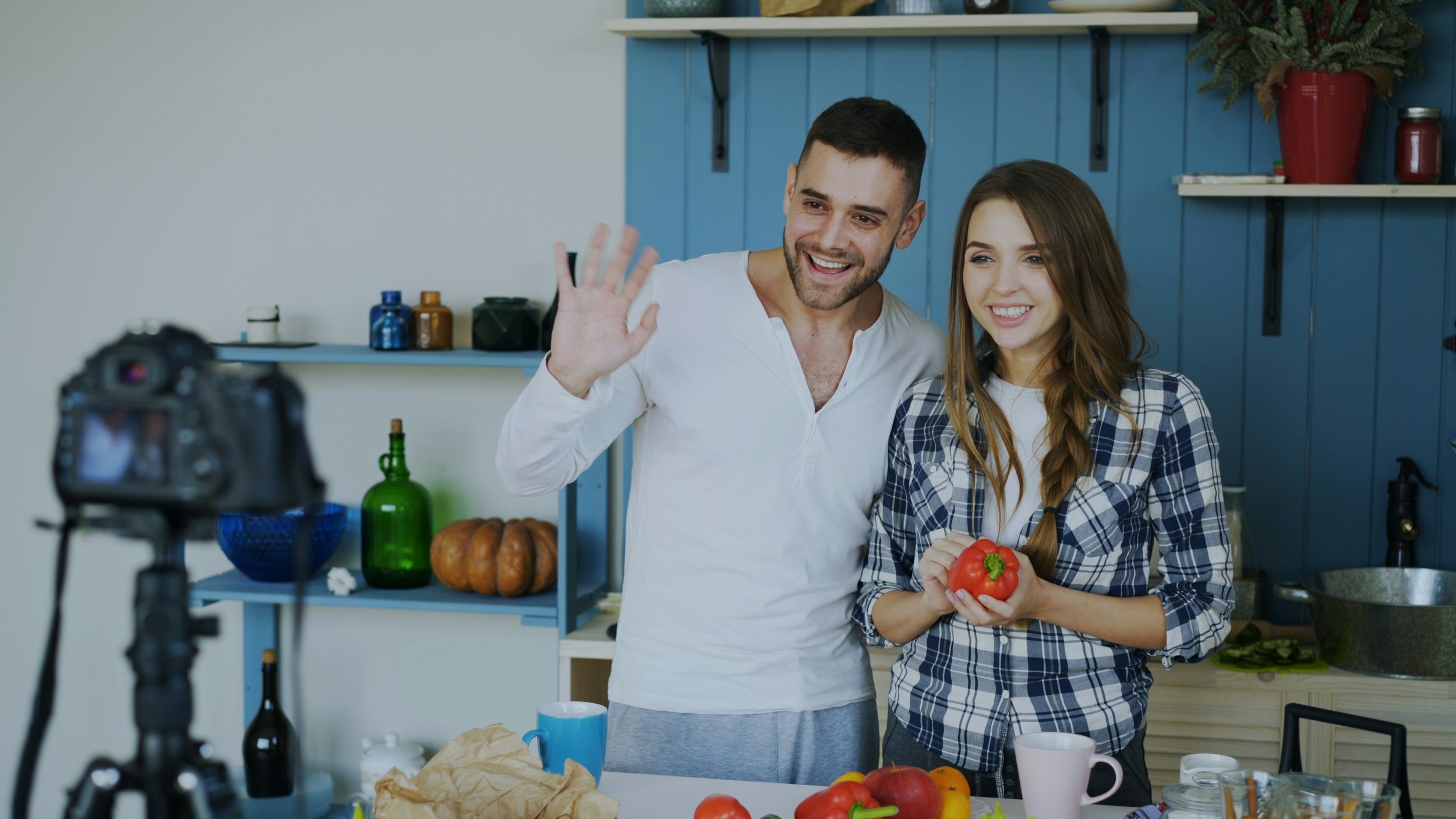 A man and woman standing in a kitchen, smiling and waving at a camera while holding a red bell pepper. The kitchen has a blue wooden wall with shelves holding plants, jars, and kitchen items. The countertop is cluttered with various fruits and vegetables, cups, and paper.