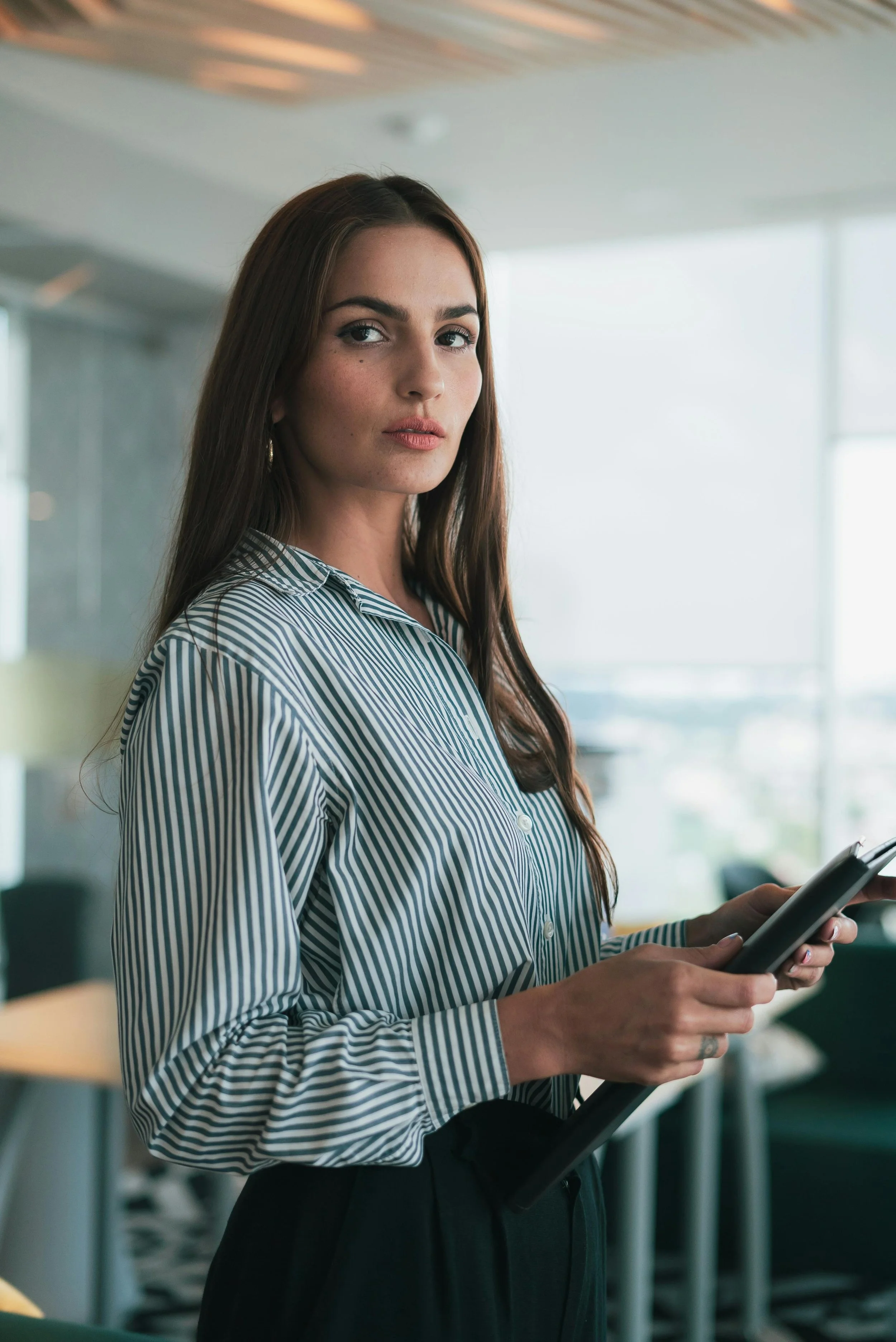 Femme debout dans un bureau moderne tenant une tablette ou un téléphone, portant une chemise à rayures.