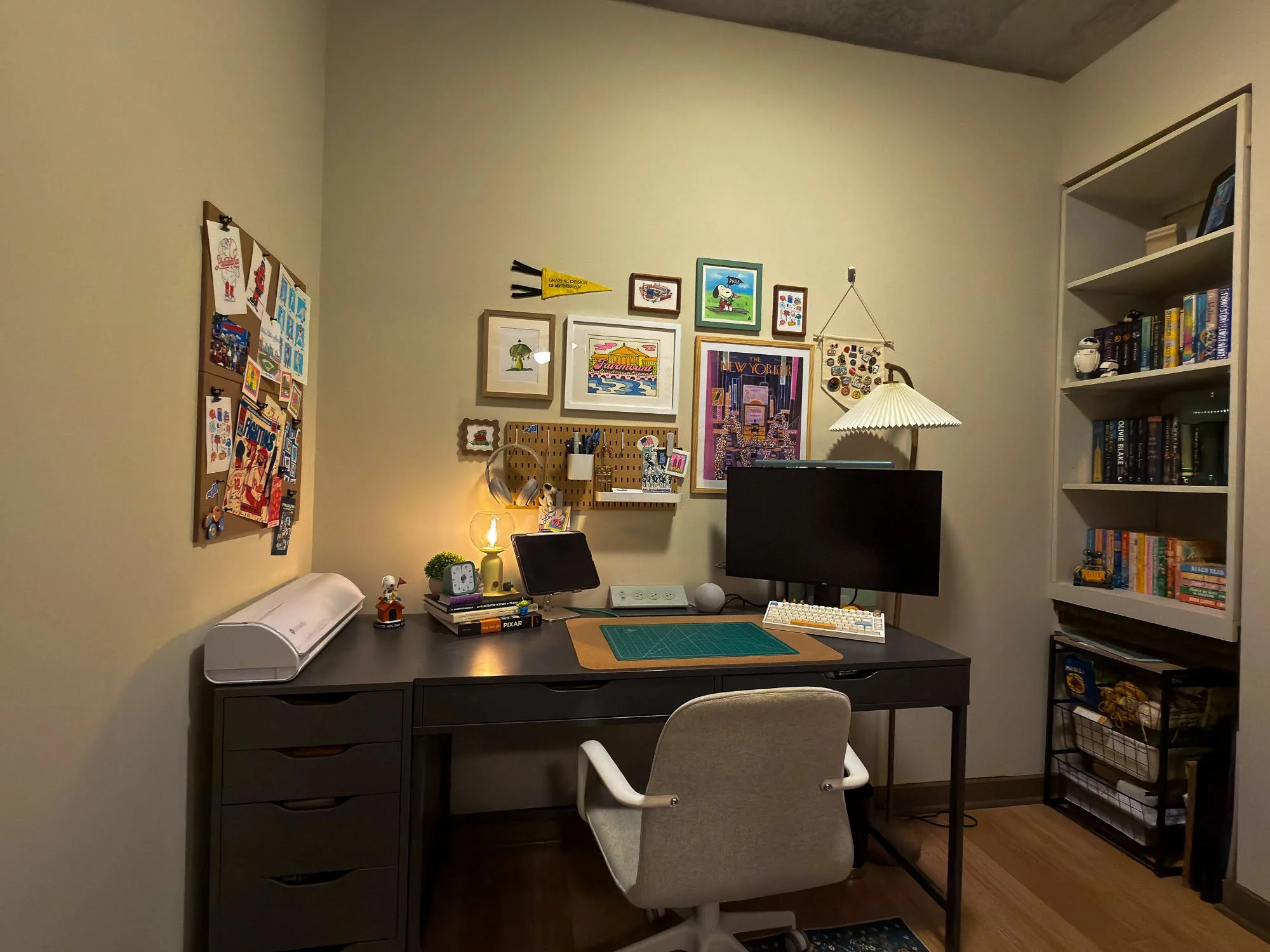 A home office with a black desk, white swivel chair, bookshelf, and wall decorations. Office supplies, a lamp, and decor items on the desk and shelves.