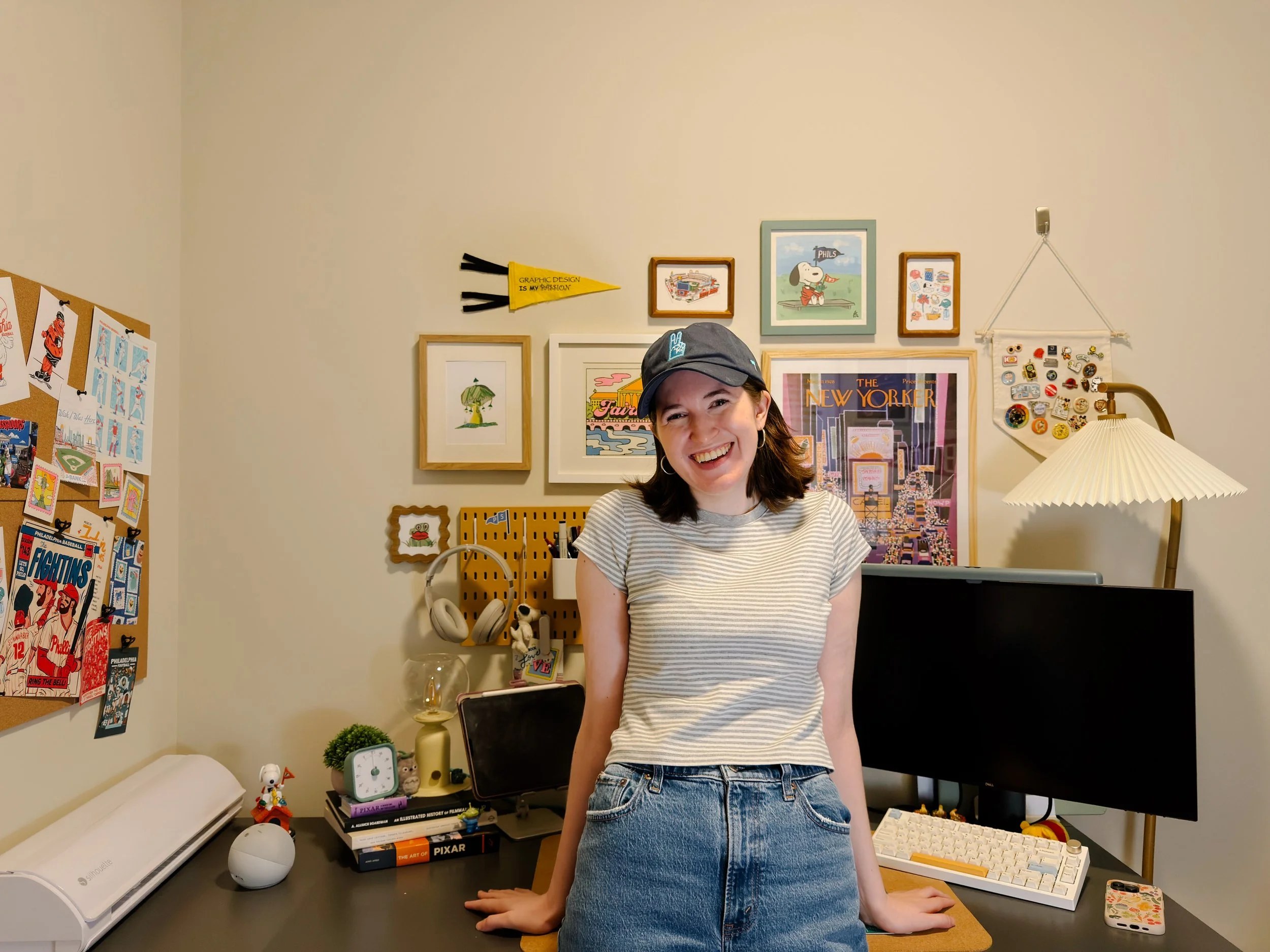 A woman smiling, wearing a striped shirt, jeans, and a dark baseball cap, standing in a cozy home office with framed artwork, a memo board with pins, books, a computer monitor, and a lamp on the desk.