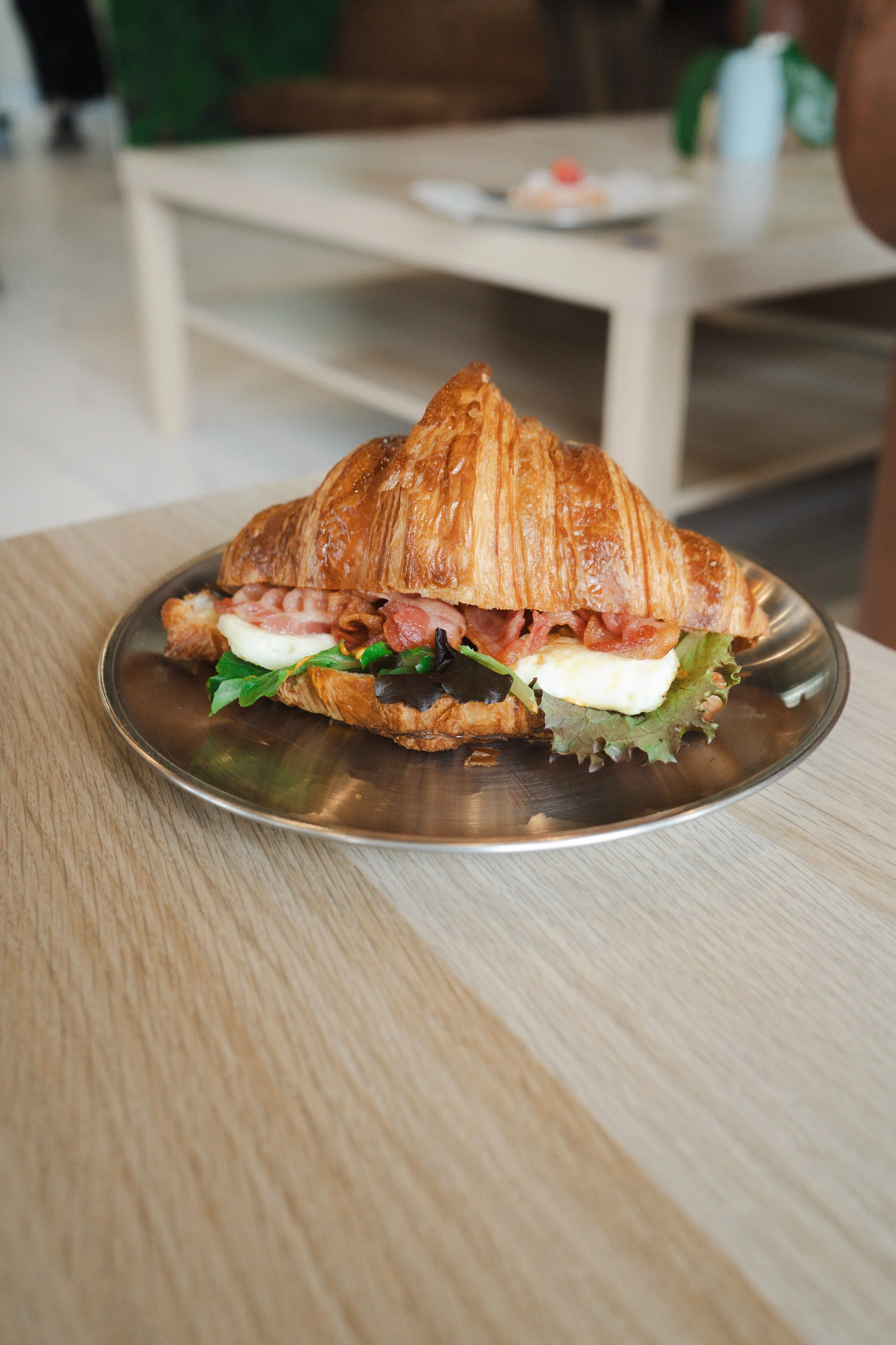 A croissant sandwich on a metal plate with bacon, lettuce, tomato, and a fried egg. In the background, there is a white shelf, a plate with a pastry, and a plant in a white vase.