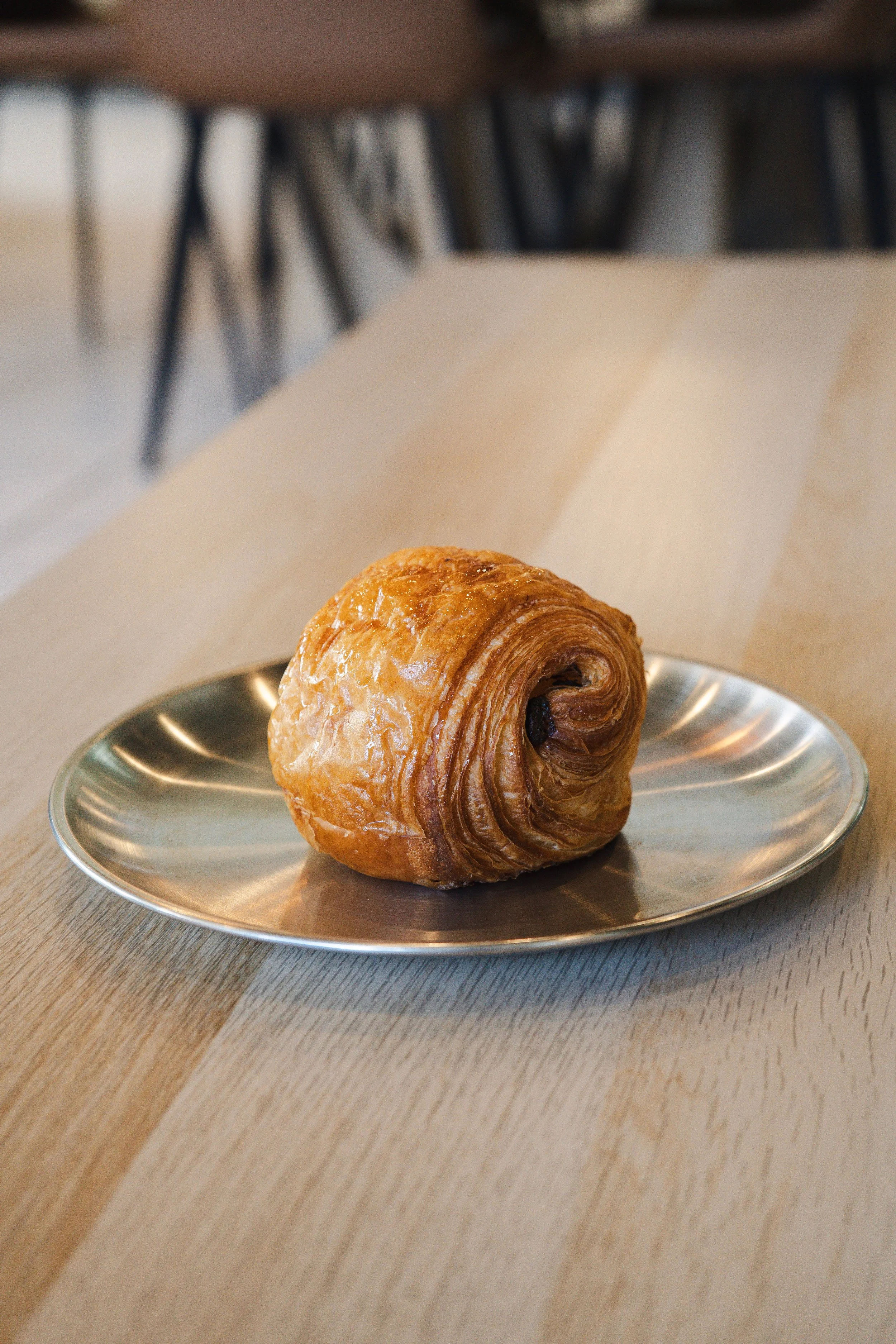 A golden brown, flaky croissant with chocolate filling on a reflective silver plate, placed on a wooden table with a background featuring blurry black chair legs and a second table.