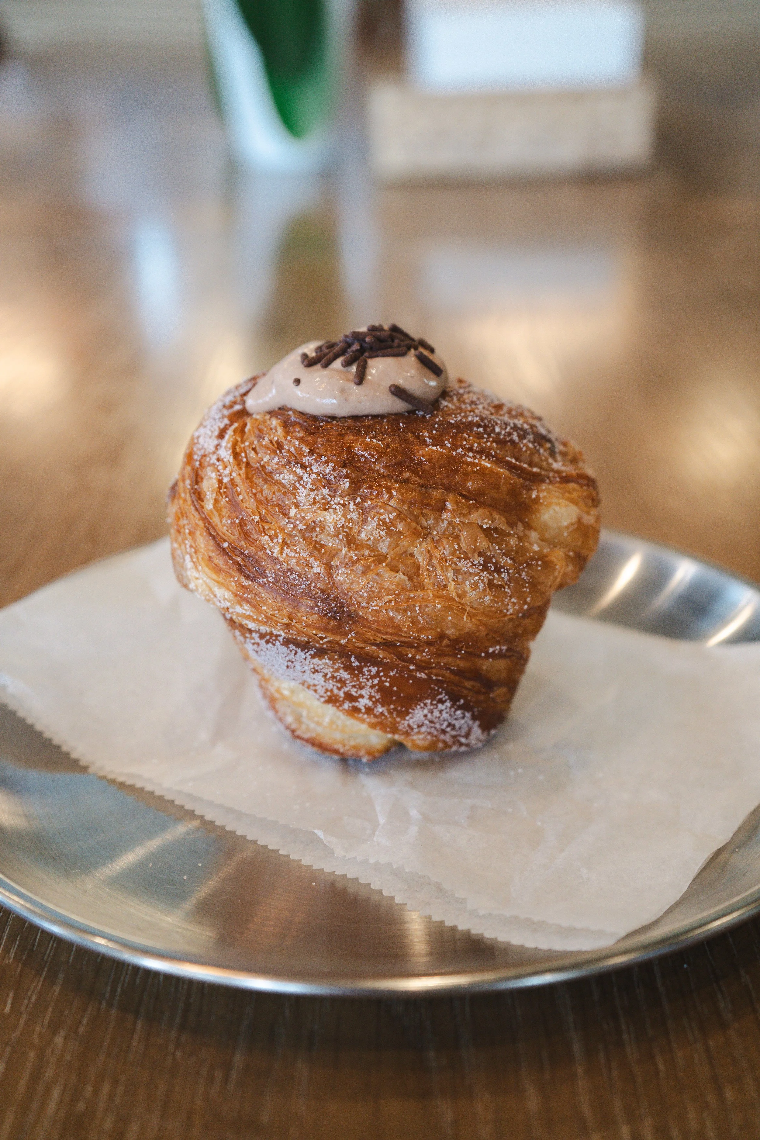 A pastry with a swirl shape, topped with a dollop of cream and chocolate sprinkles, dusted with powdered sugar, on a piece of parchment paper in a metal serving dish.