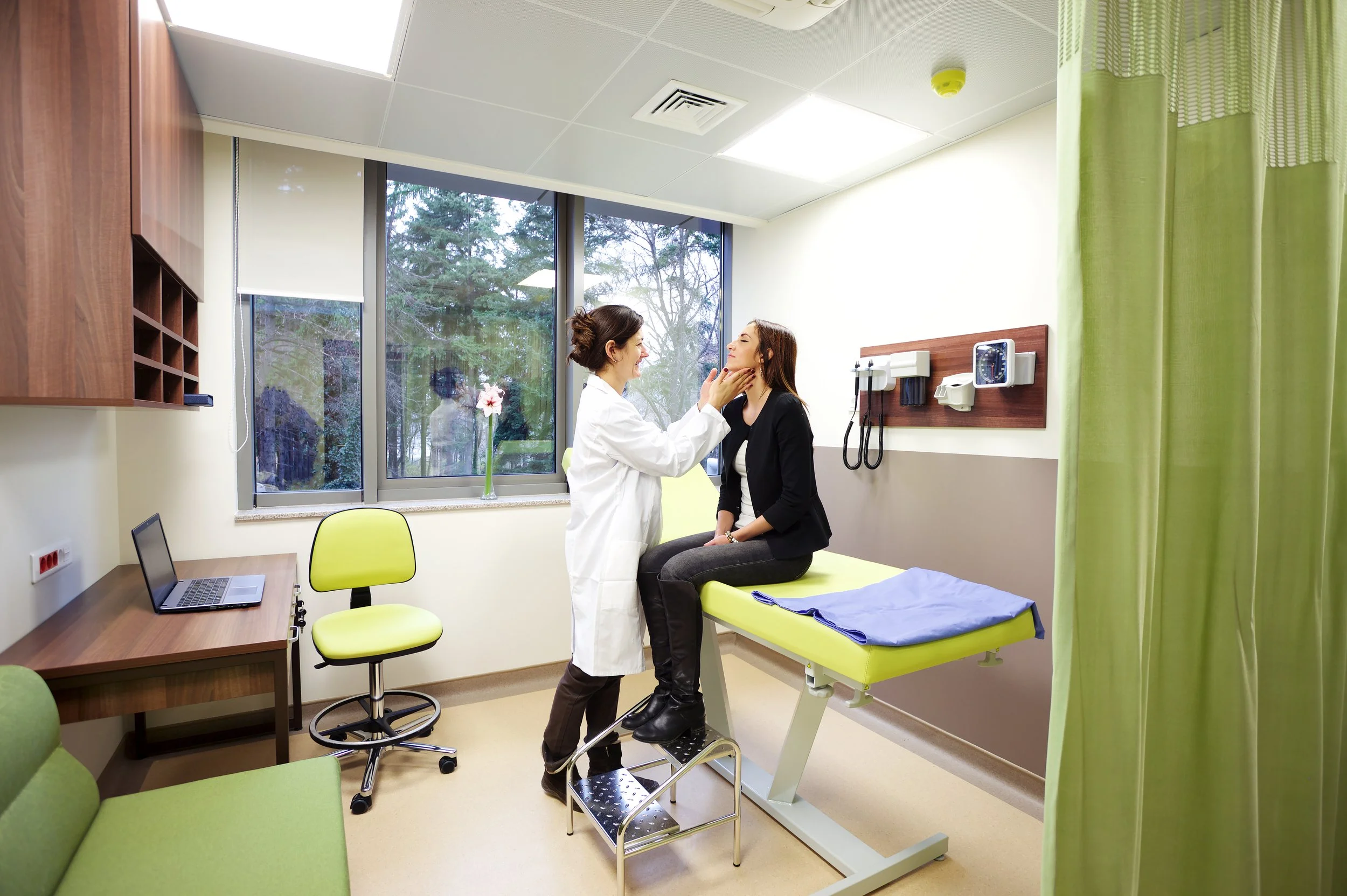 A healthcare professional, wearing a white coat, examines a patient sitting on an examination table in a medical room with a large window and medical equipment on the wall.