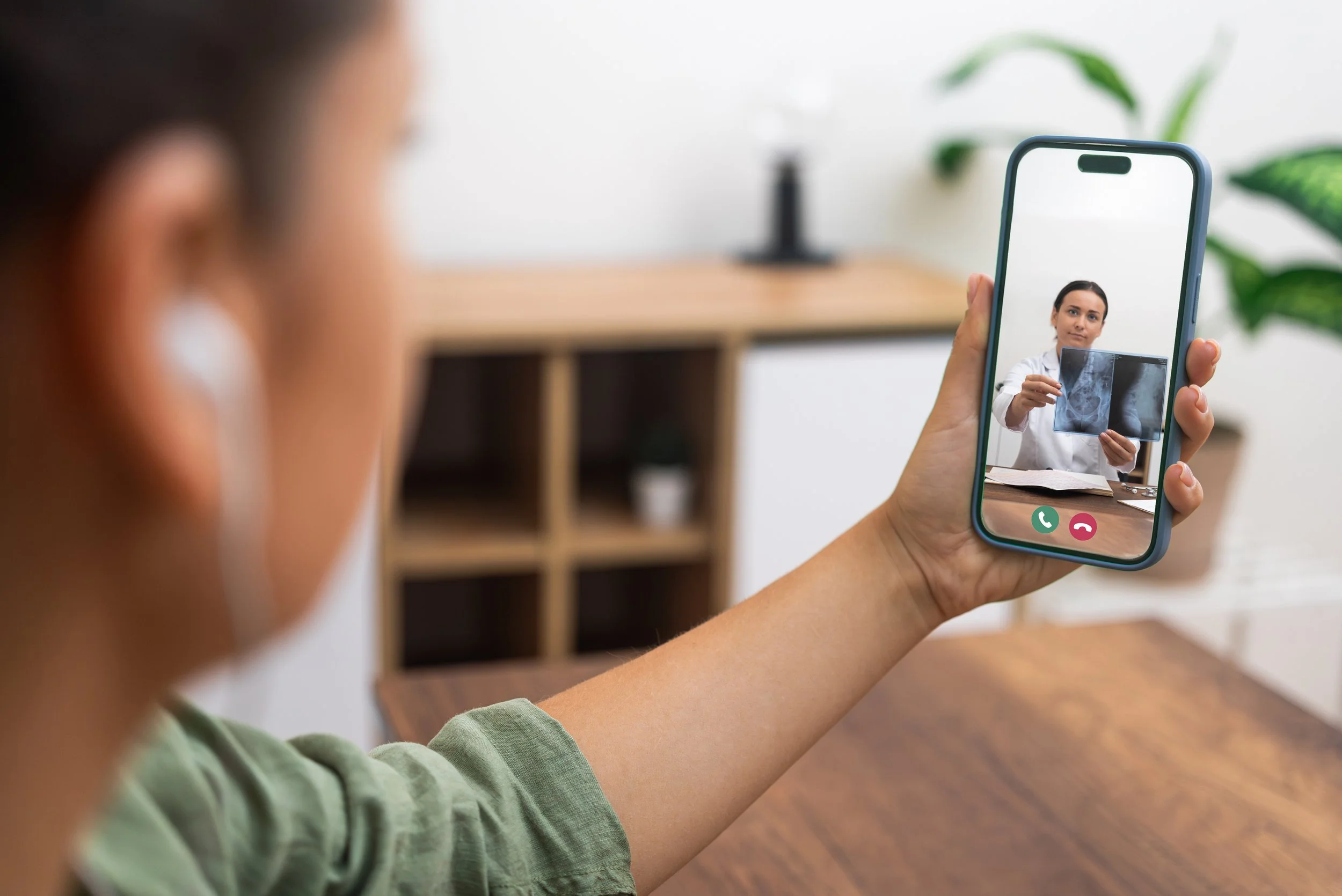 A woman using her phone, on a video call with her GP as the doctor on the screen sits in their office holding a chest X-ray to the screen.