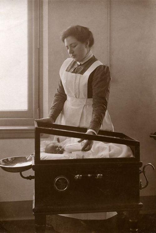 A midwife in vintage uniform looking at a newborn baby in a hospital incubator.