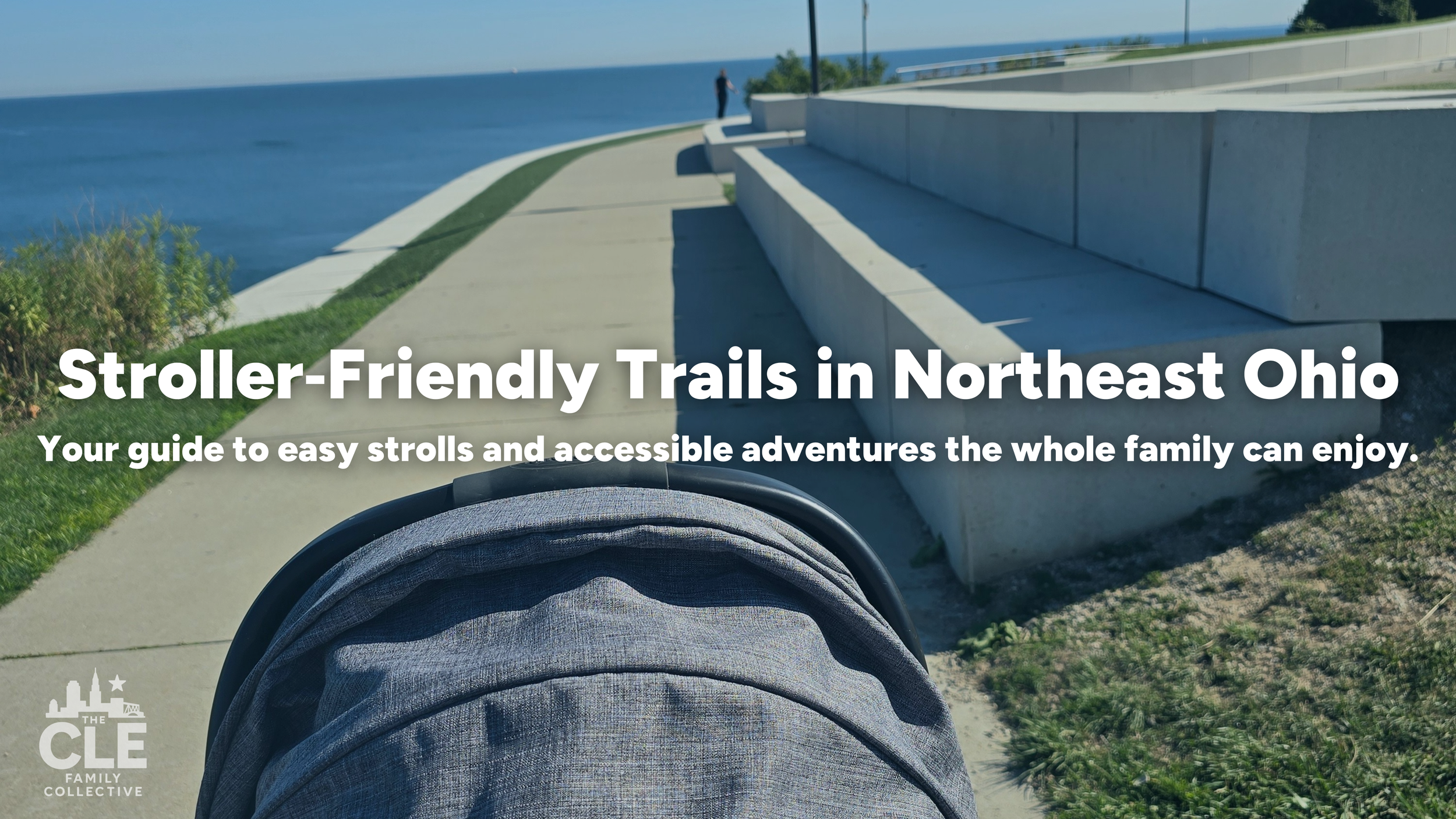 A stroller on a paved trail along a body of water with a person standing in the distance. Sunlight and clear weather are visible.