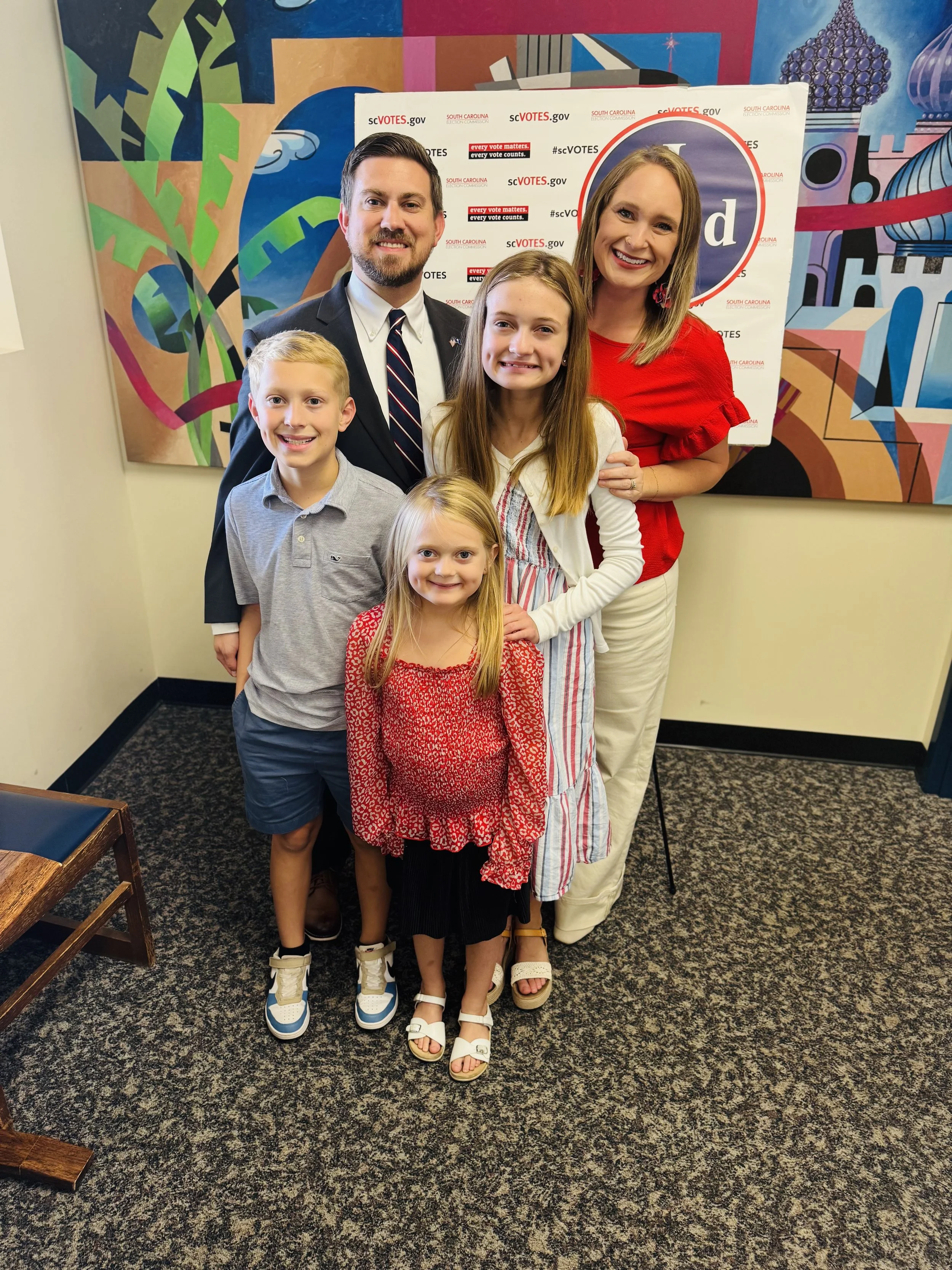 Family standing together at a voting event, with a colorful mural in the background and a South Carolina election sign.